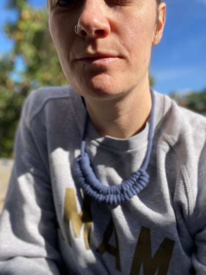 Person wearing a grey sweatshirt & a blue necklace, sitting outdoors with greenery in the background.