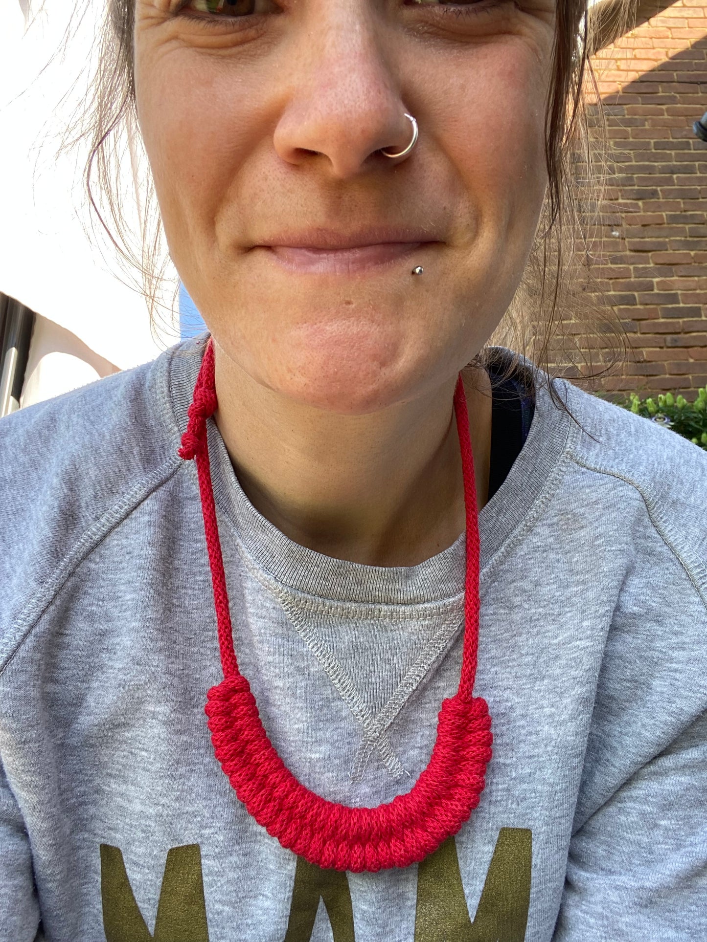 Person smiling at the camera, wearing a grey sweatshirt with a red necklace, outdoors.
