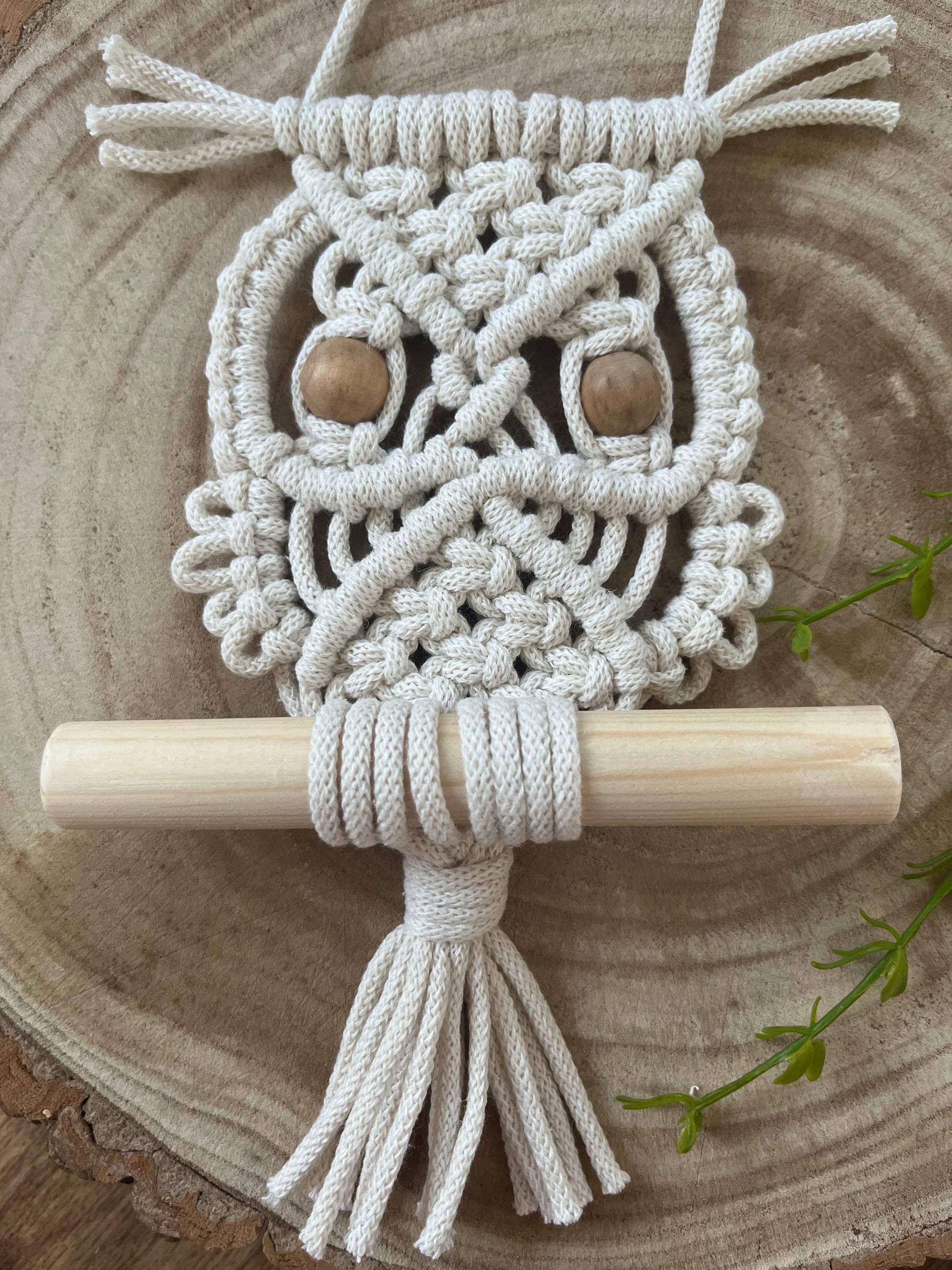 Close up of a handmade macrame owl wall hanging decoration, perched on a wooden dowel. Made with natural cotton and wooden beads, displayed on a wooden board.  