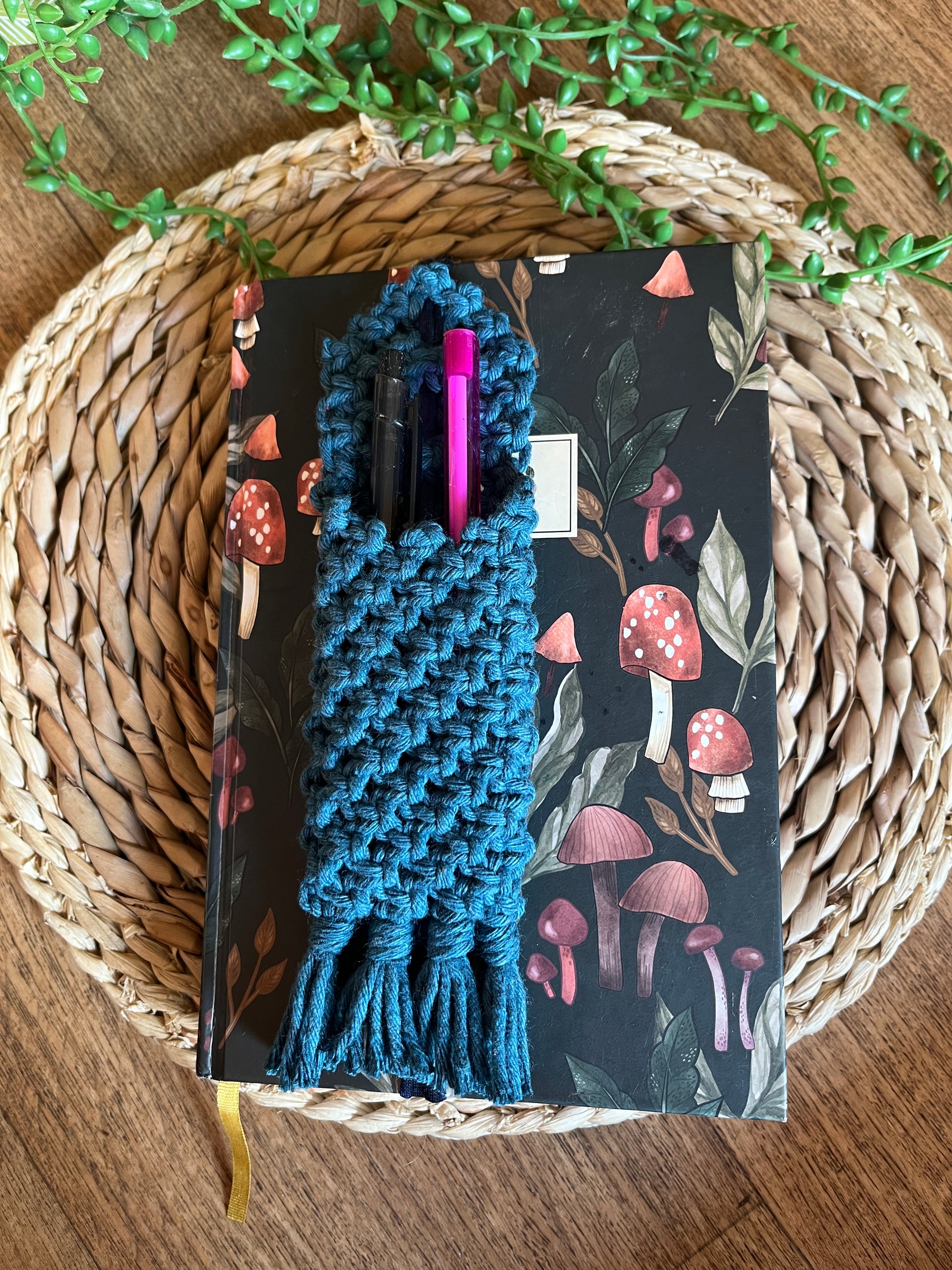Handmade macrame wrap around pen holder, made with petrol blue recycled cotton yarn. Displayed on a notebook which has a mushroom pattern, on a wicker mat. 