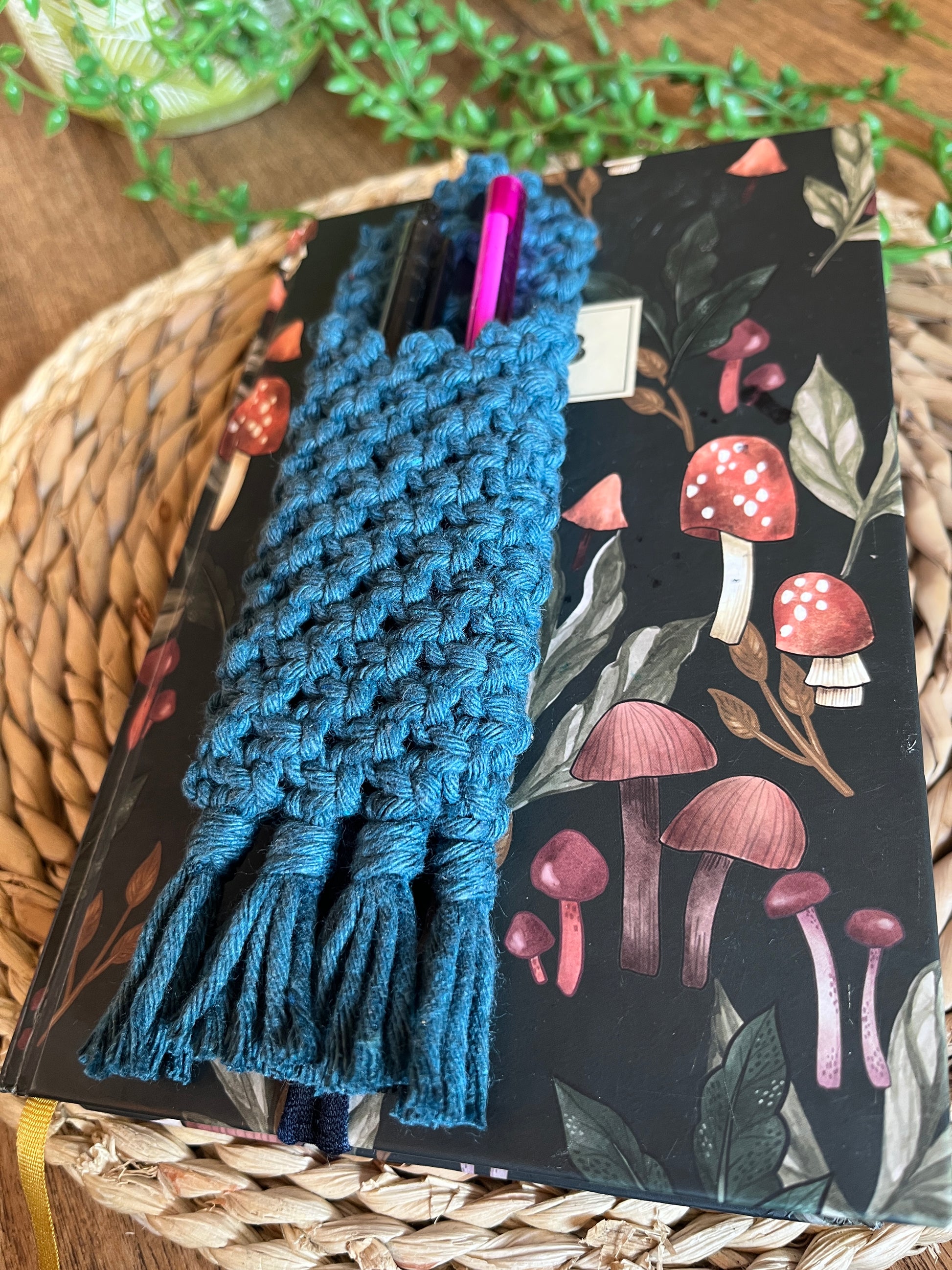 Handmade macrame wrap around pen holder, made with petrol blue recycled cotton yarn. Displayed on a notebook which has a mushroom pattern, on a wicker mat. 