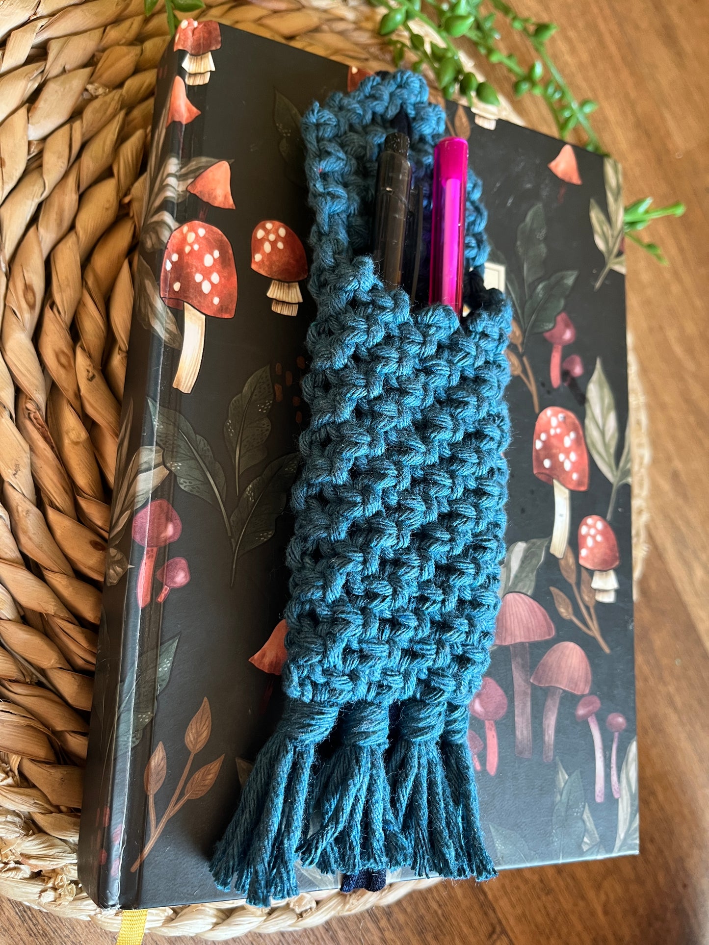 Handmade macrame wrap around pen holder, made with petrol blue recycled cotton yarn. Displayed on a notebook which has a mushroom pattern, on a wicker mat. 