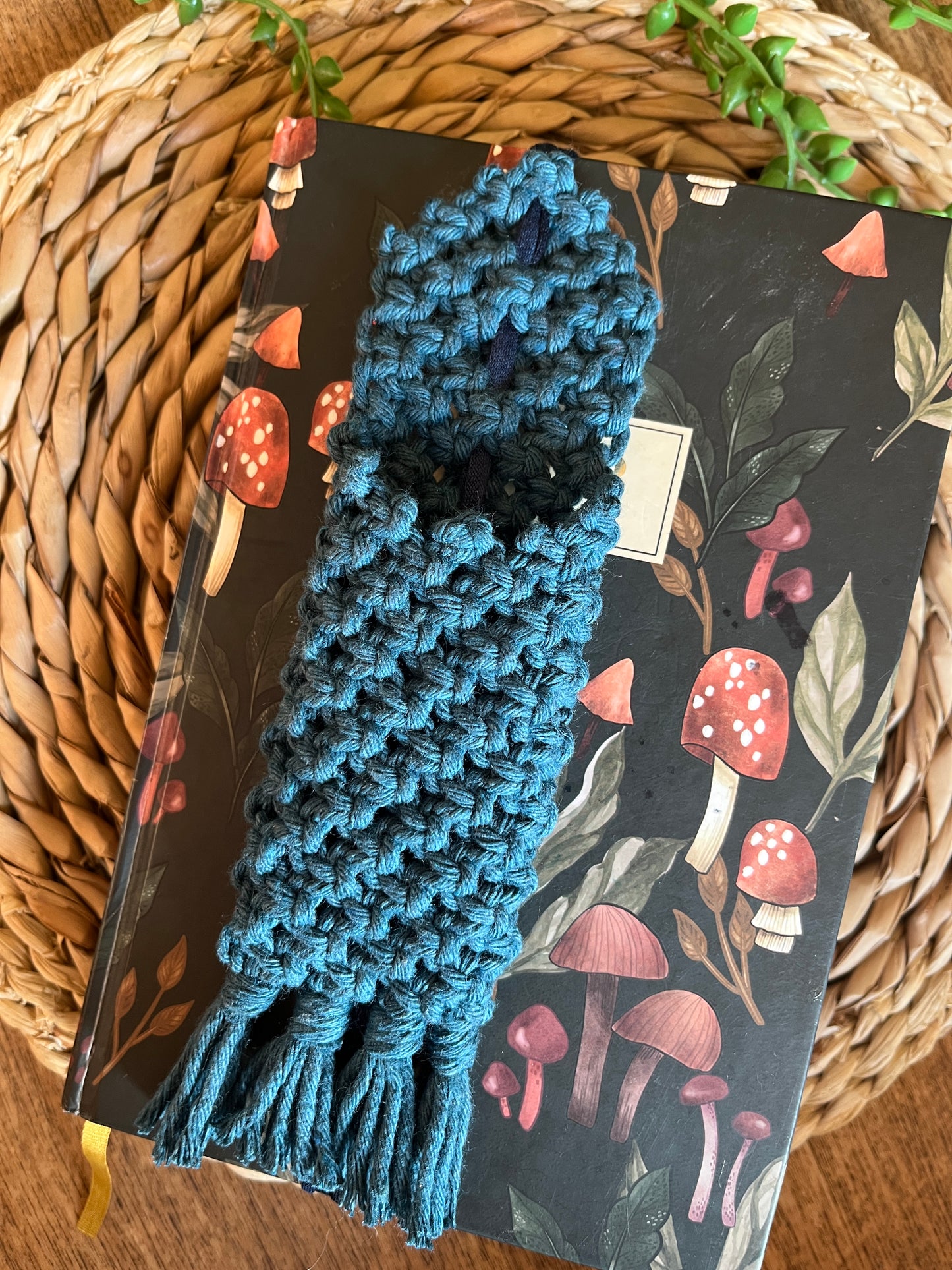 Handmade macrame wrap around pen holder, made with petrol blue recycled cotton yarn. Displayed on a notebook which has a mushroom pattern, on a wicker mat. 