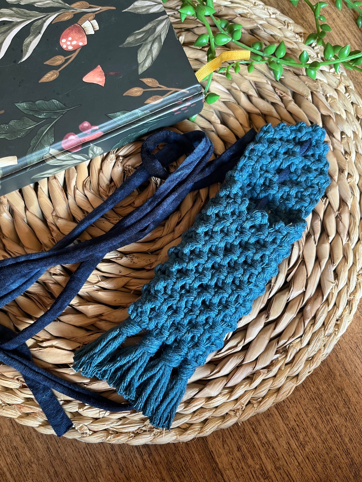 Handmade macrame wrap around pen holder, made with petrol blue recycled cotton yarn. Displayed next to a notebook which has a mushroom pattern, on a wicker mat. 