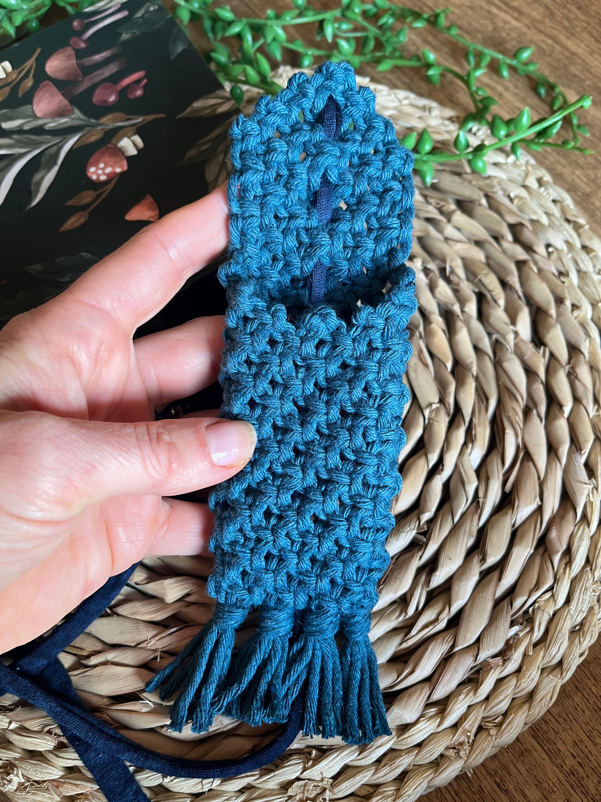 A hand holding a handmade macrame wrap around pen holder, made with petrol blue recycled cotton yarn. Displayed next a notebook which has a mushroom pattern, on a wicker mat. 