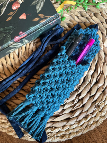 Handmade macrame wrap around pen holder, made with petrol blue recycled cotton yarn. Displayed next a notebook which has a mushroom pattern, on a wicker mat. The pen holder has two pens inside of it, one is black, one is pink.