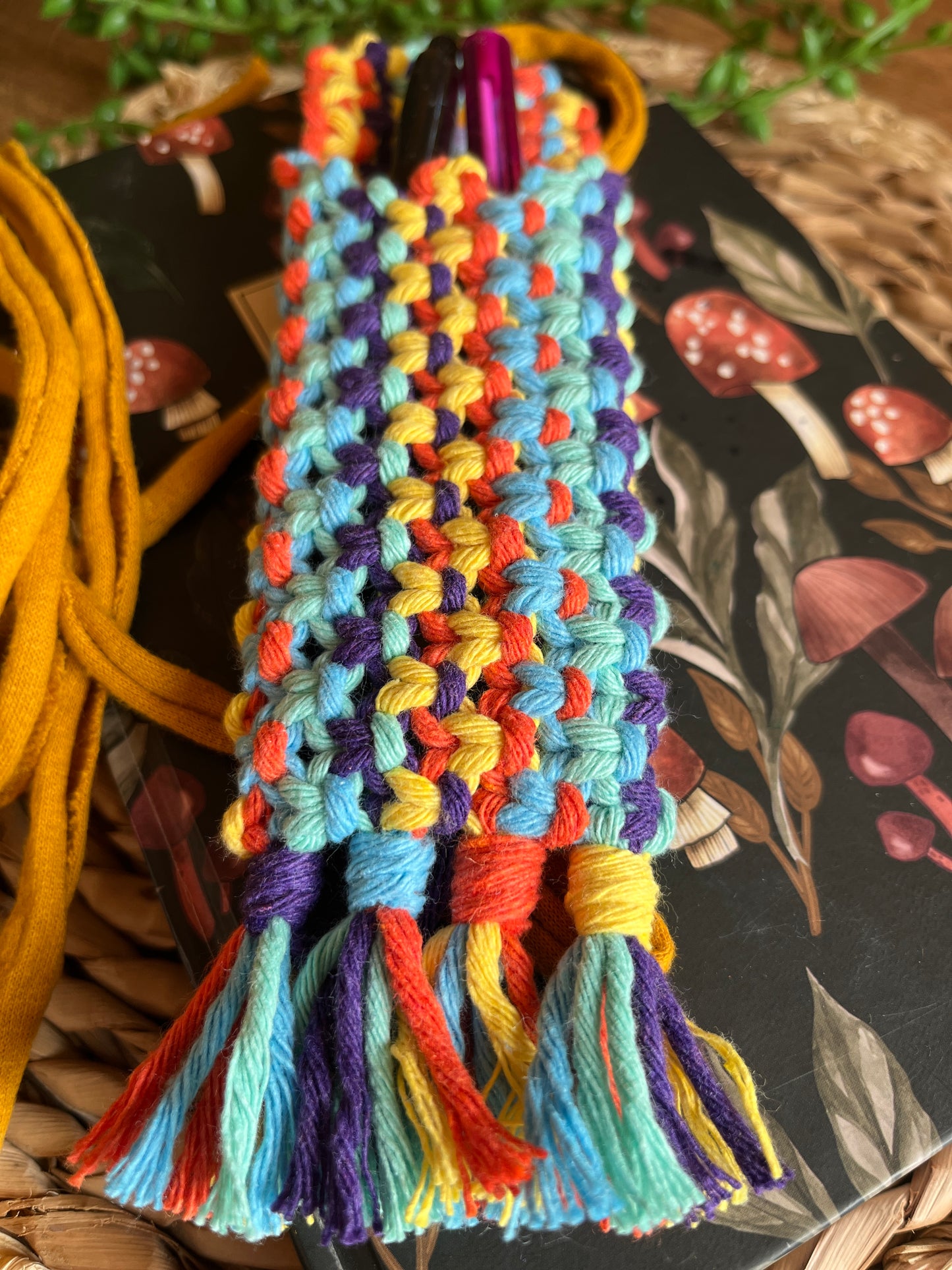 Handmade macrame wrap around pen holder, made with multiple colours of recycled cotton yarn. Displayed on a notebook which has a mushroom pattern, on a wicker mat. The pen holder has two pens inside of it, one black and one pink.