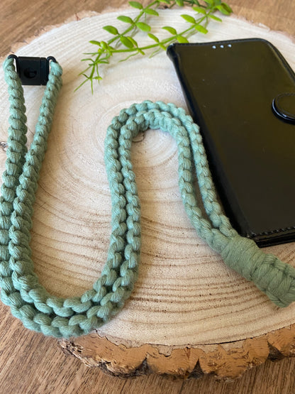 A black leather phone case on a wooden board with a macrame phone lanyard made with green yarn attached to it with a metal clasp. 