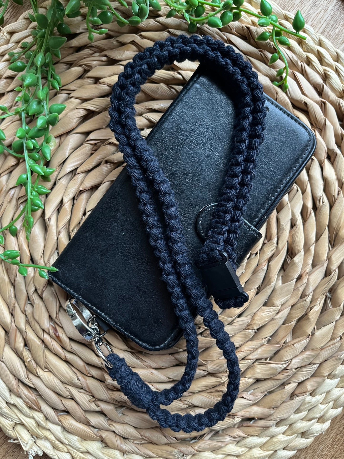 A black phone case on a wicker mat with a macrame phone lanyard made with navy blue yarn attached to it with a metal clasp. 
