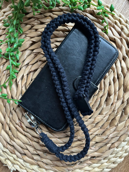 A black phone case on a wicker mat with a macrame phone lanyard made with navy blue yarn attached to it with a metal clasp. 