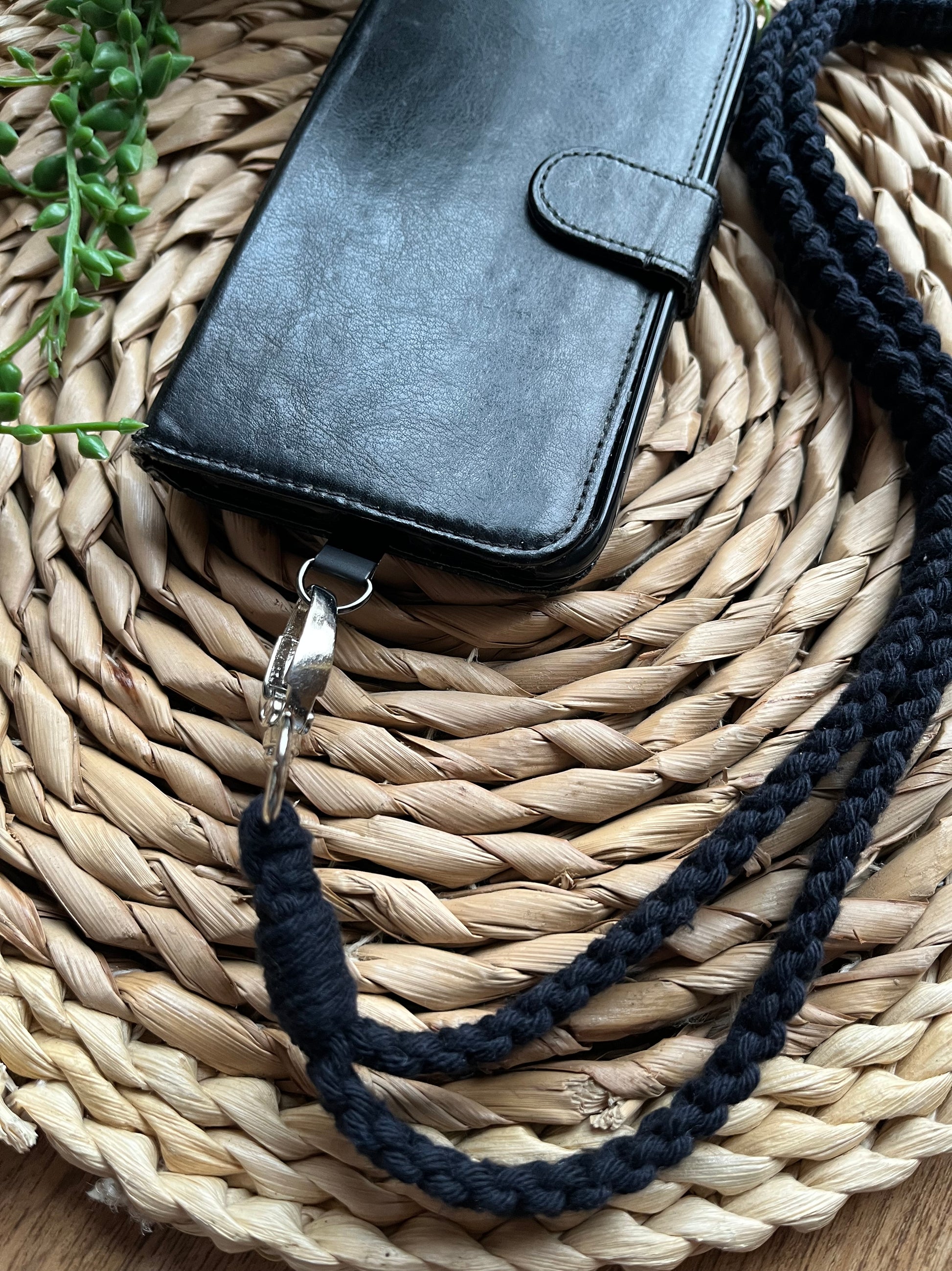 Close up of a black phone case on a wicker mat with a macrame phone lanyard made with navy blue yarn attached to it with a metal clasp. 