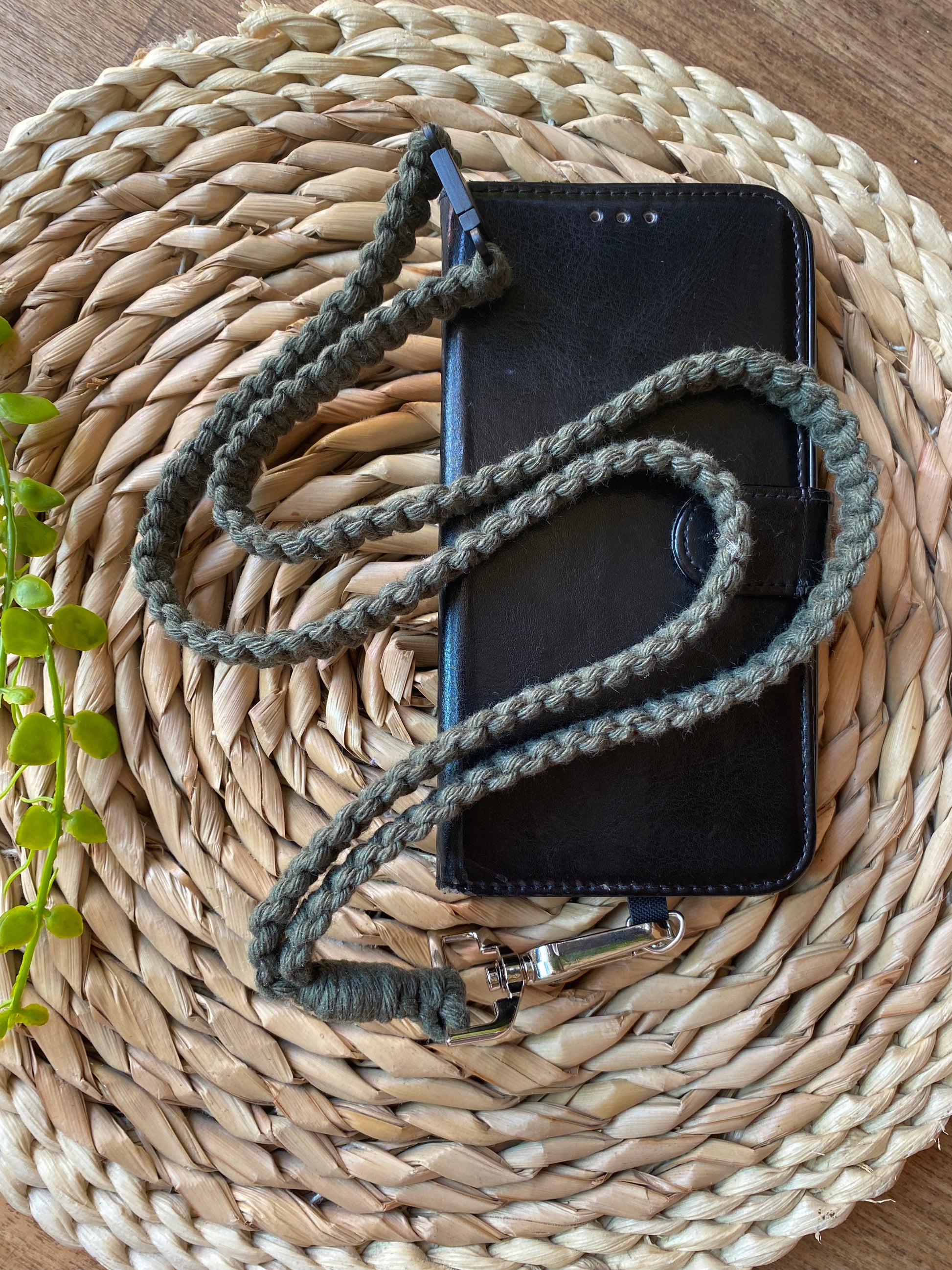 A black leather phone case on a wooden board with a macrame phone lanyard made with olive green yarn attached to it with a metal clasp. 