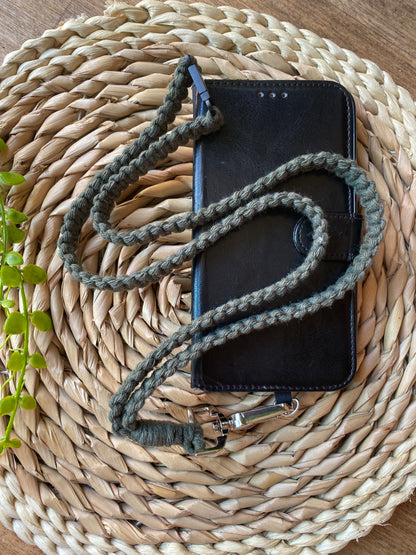 A black leather phone case on a wooden board with a macrame phone lanyard made with olive green yarn attached to it with a metal clasp. 