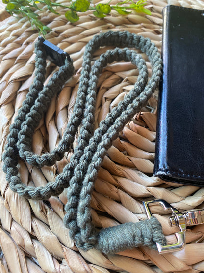 Close up of a black leather phone case on a wicker placemat with a macrame phone lanyard made with olive green yarn attached to it with a metal clasp. 