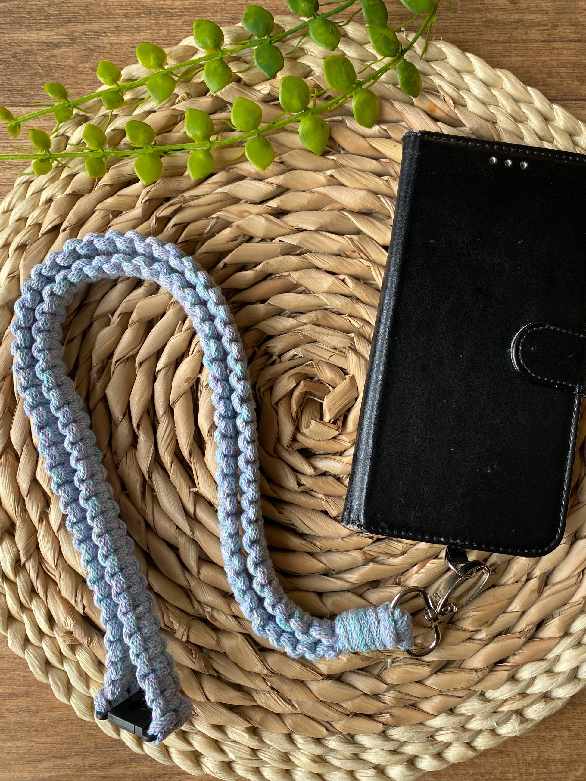 A black leather phone case on a wicker mat with a macrame phone lanyard made with pastel coloured yarn attached to it with a metal clasp. 