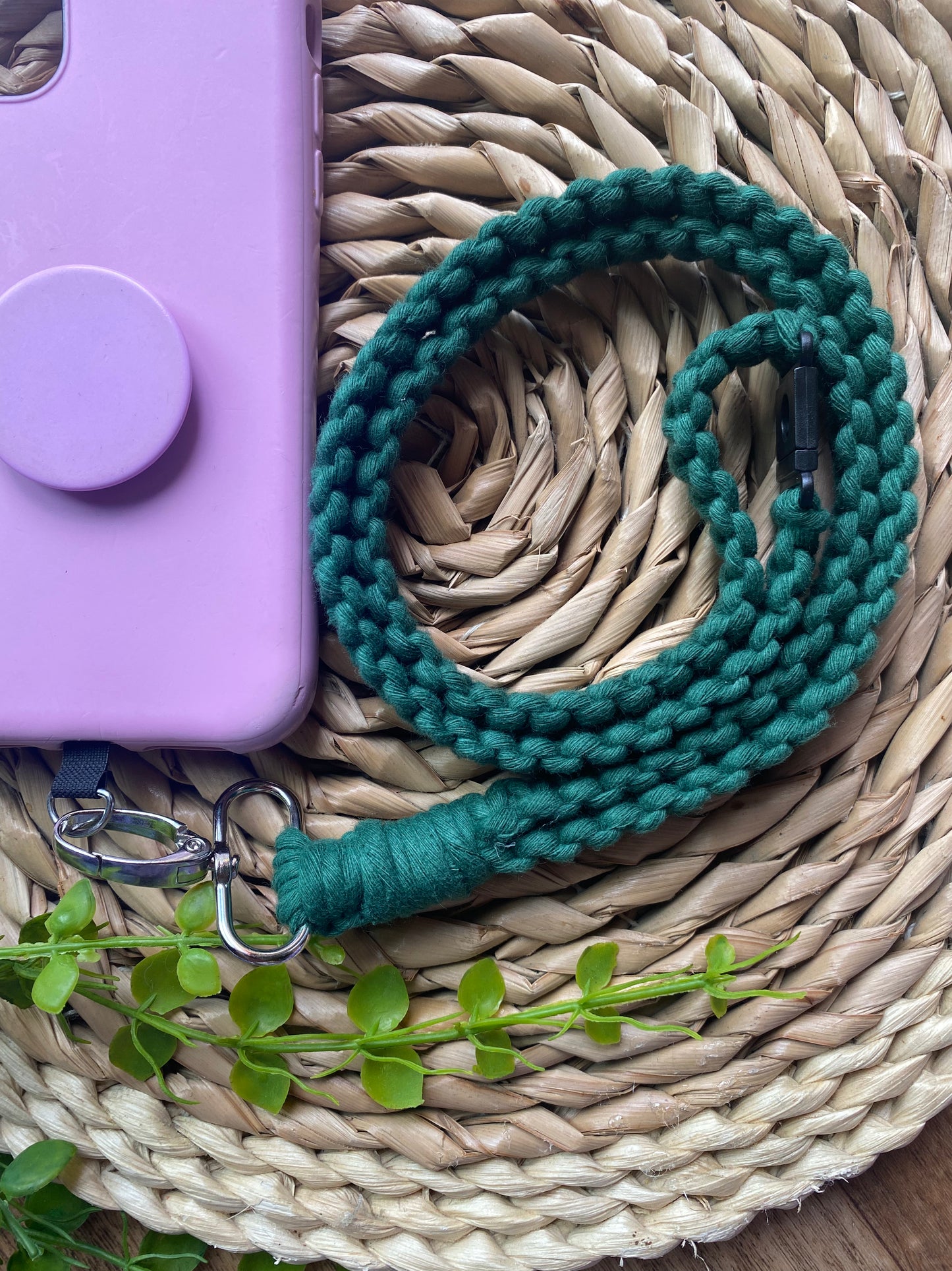 A pink phone case on a wicker mat with a macrame phone lanyard made with pine green yarn attached to it with a metal clasp. 