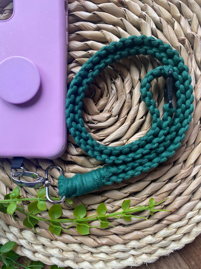 A pink phone case on a wicker mat with a macrame phone lanyard made with pine green yarn attached to it with a metal clasp. 