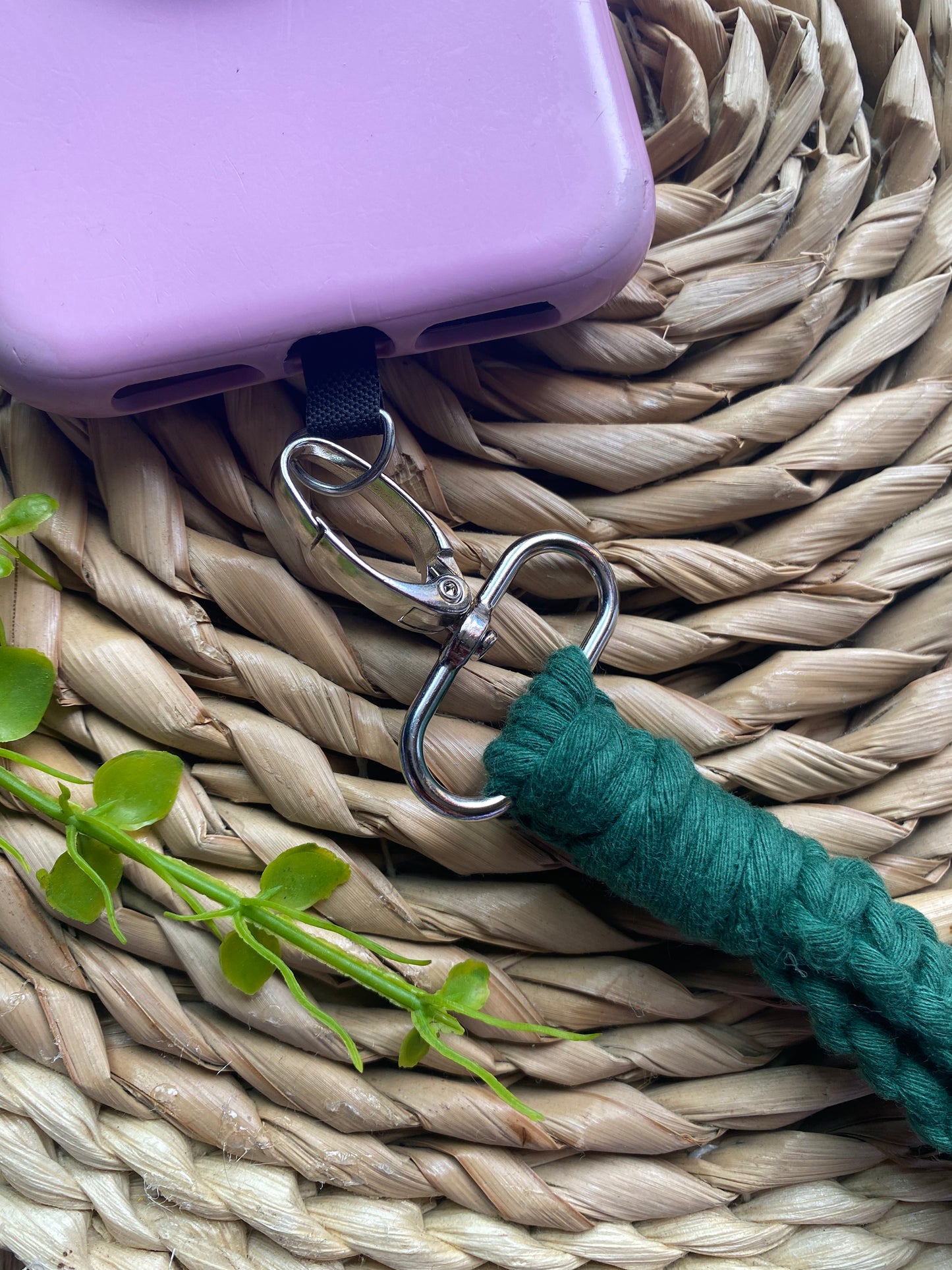 Close up of a pink phone case on a wicker mat with a macrame phone lanyard made with pine green yarn attached to it with a metal clasp. 