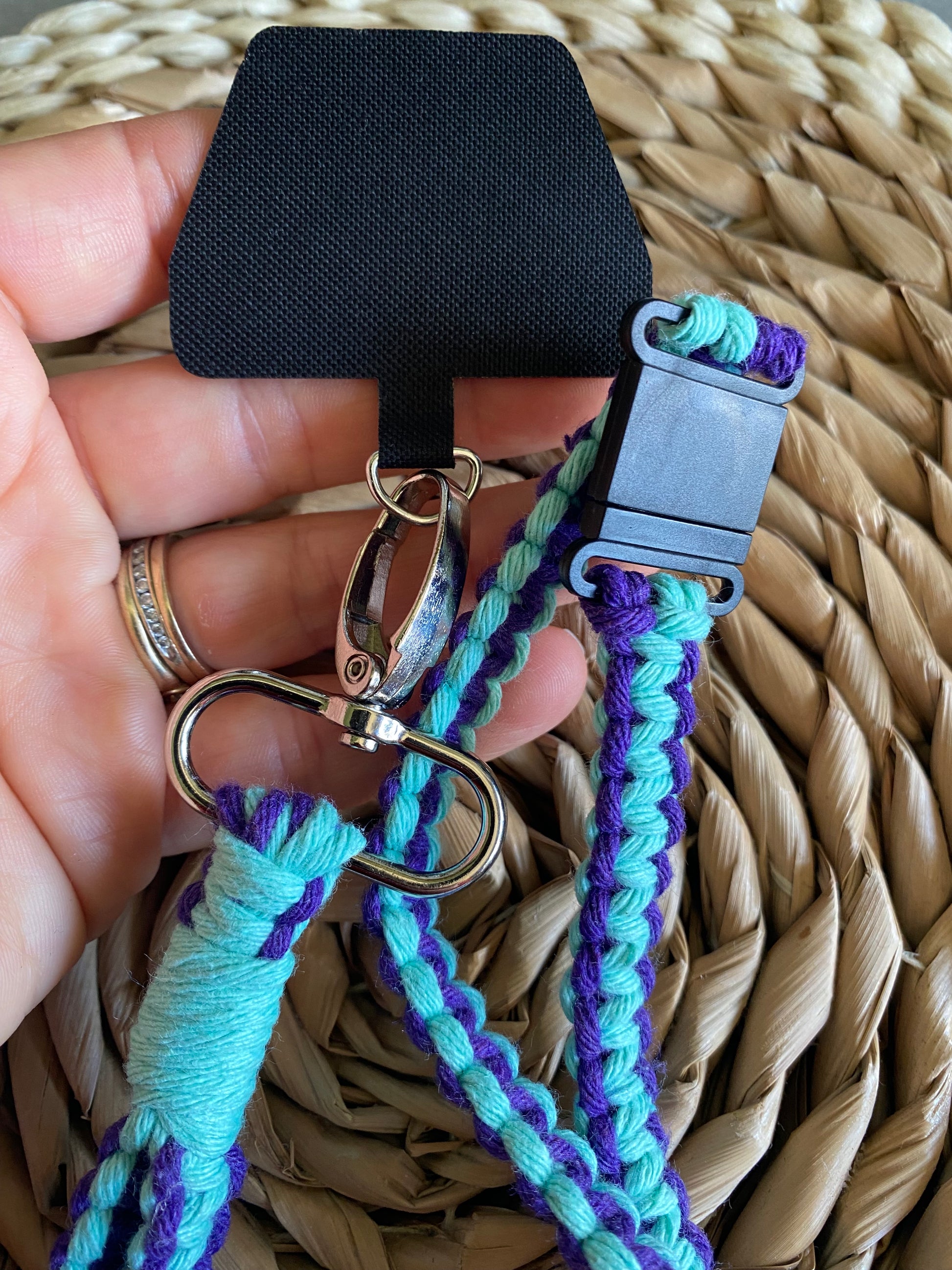 A hand holding a universal phone tether on the end of a macrame phone lanyard made with purple and mint coloured yarn and a metal clasp. 