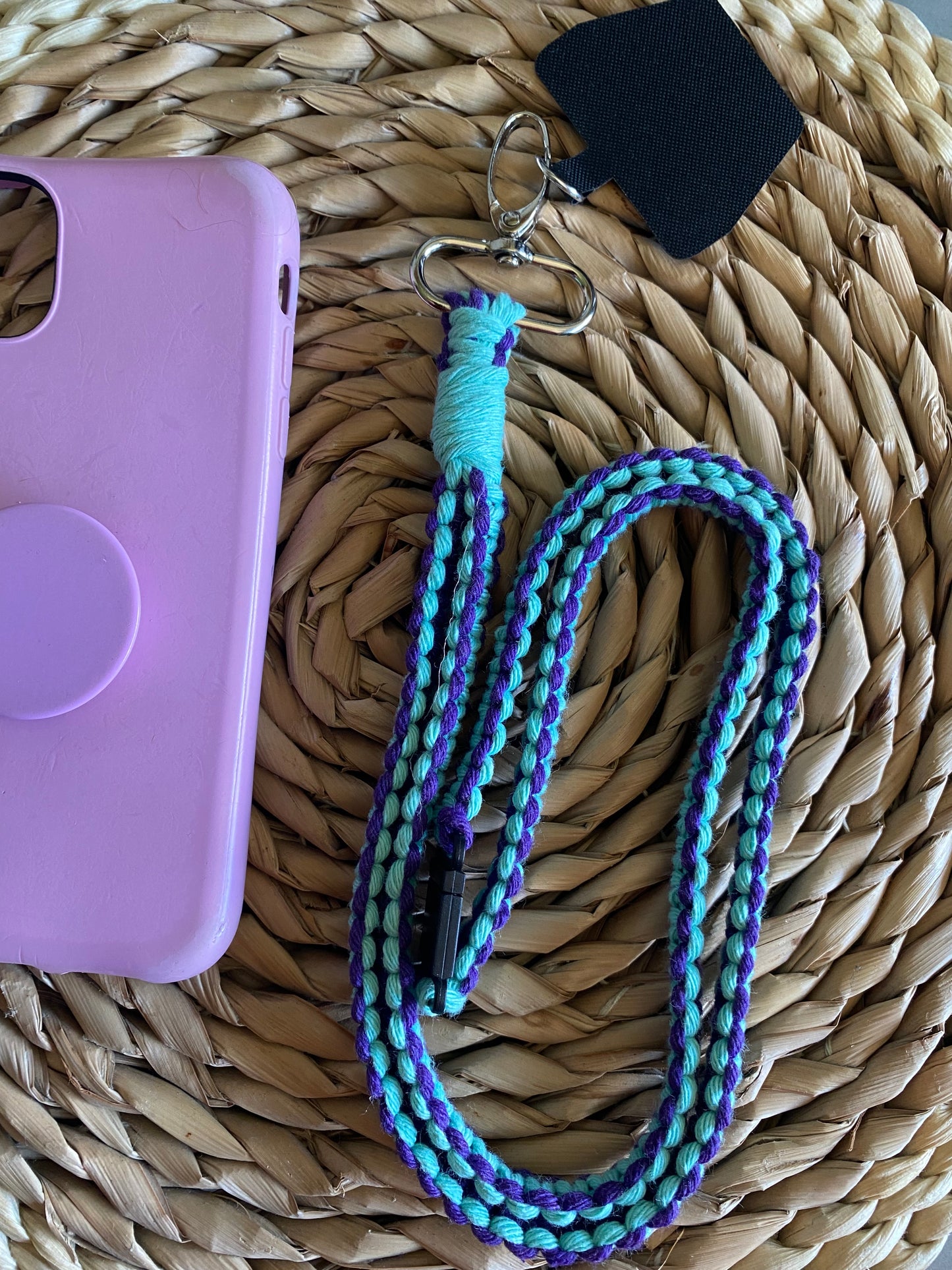 A pink phone case on a wicker mat with a macrame phone lanyard made with purple and mint yarn next to it. 