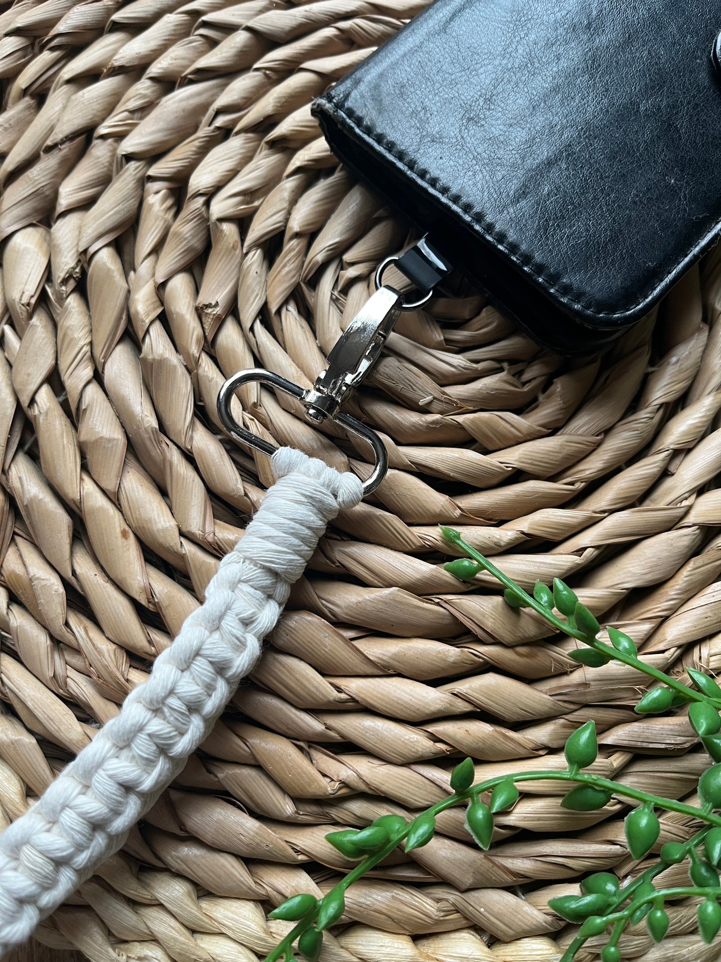White macrame strap with metal clip, attached to a black leather phone case, on a woven surface with a plant