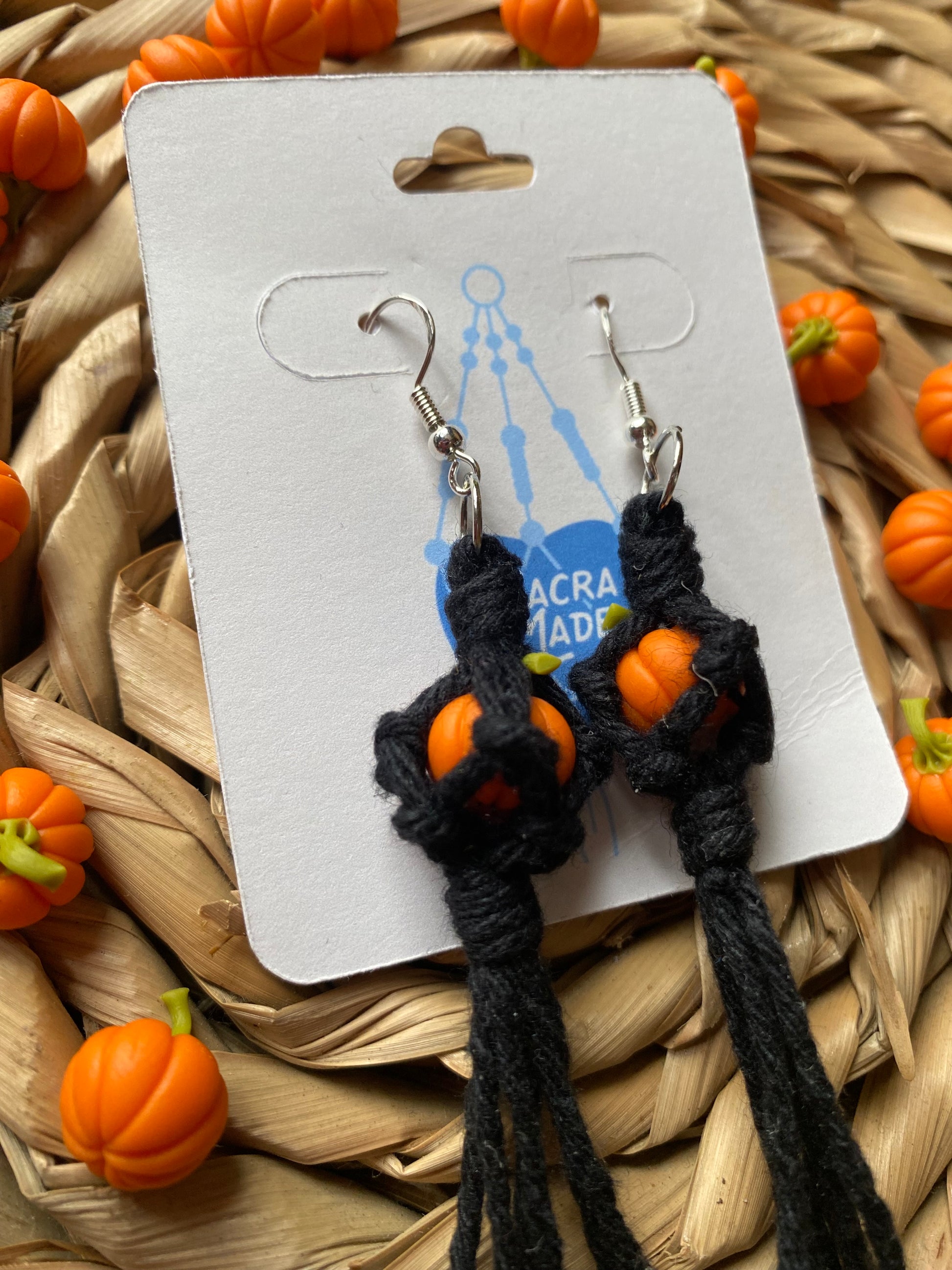 Close up of a pair of earrings made with small orange pumpkins wrapped in black yarn on silver hooks, displayed on a white backing card with a wicker mat and lots of small orange pumpkins in the background.  