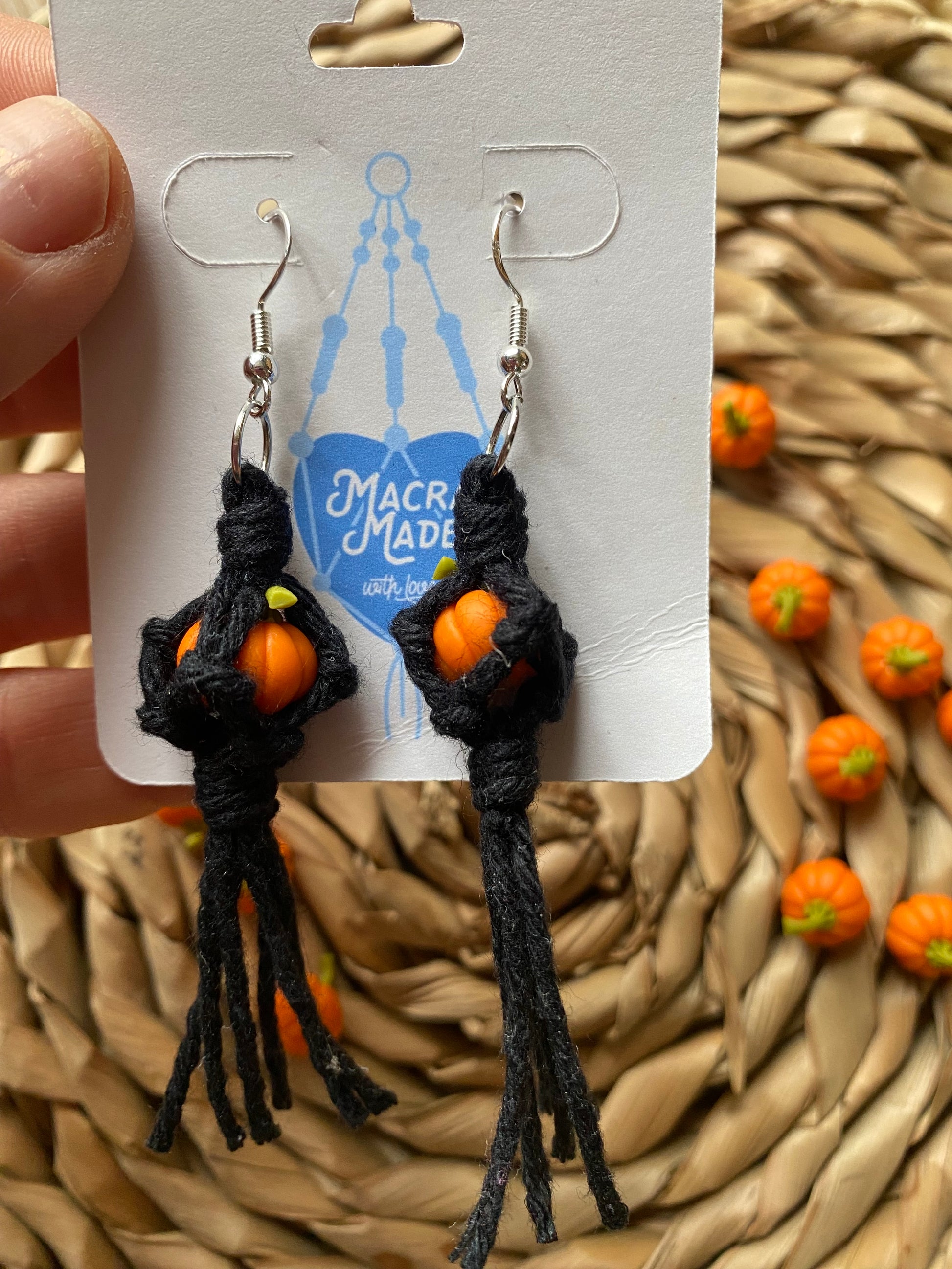 A hand holding a pair of earrings made with small orange pumpkins wrapped in black yarn on silver hooks, displayed on a white backing card with a wicker mat and lots of small orange pumpkins in the background.  