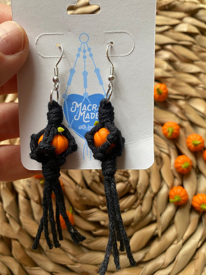 A hand holding a pair of earrings made with small orange pumpkins wrapped in black yarn on silver hooks, displayed on a white backing card with a wicker mat and lots of small orange pumpkins in the background.  