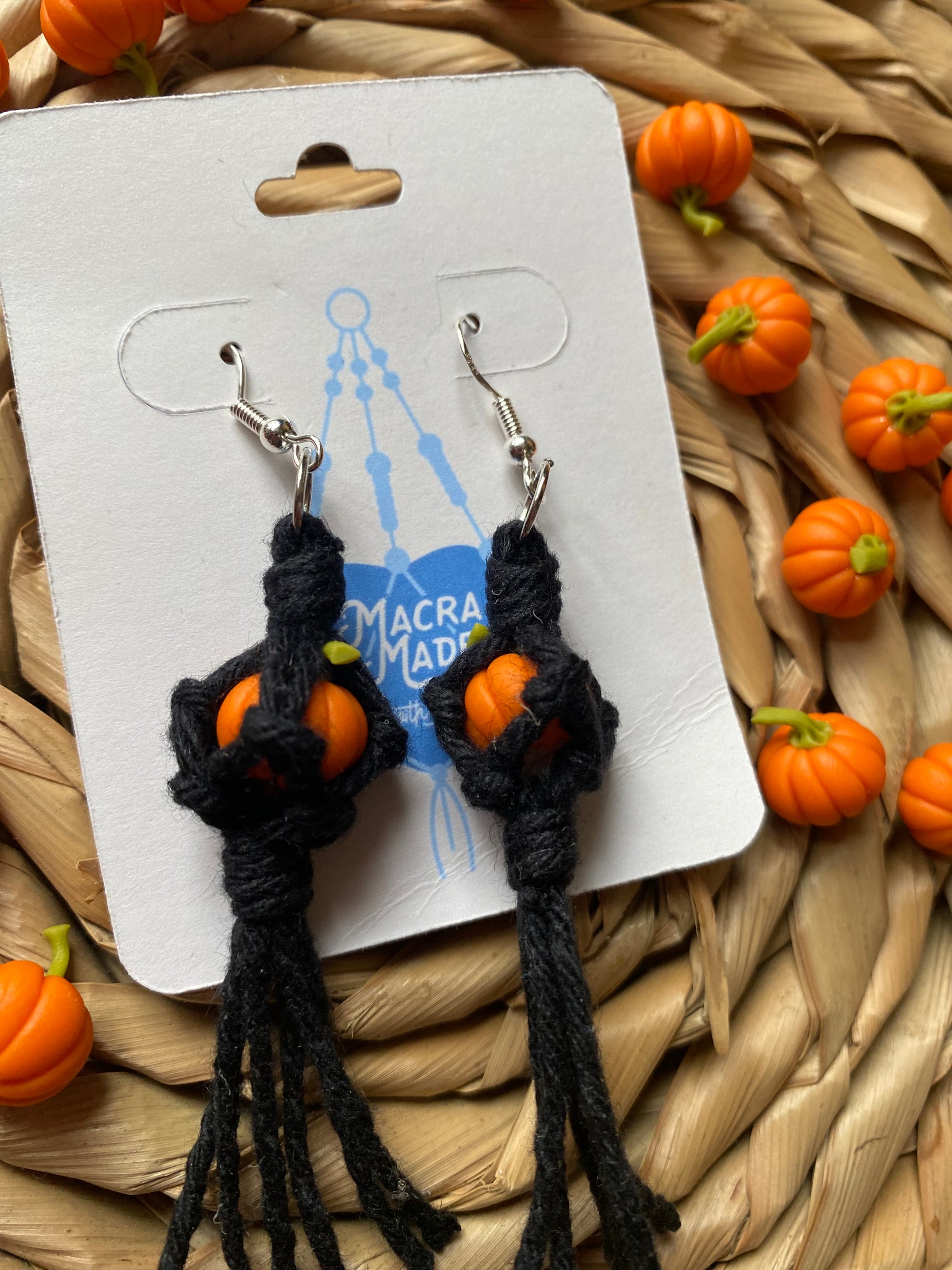 Close up of a pair of earrings made with small orange pumpkins wrapped in black yarn on silver hooks, displayed on a white backing card with a wicker mat and lots of small orange pumpkins in the background.  