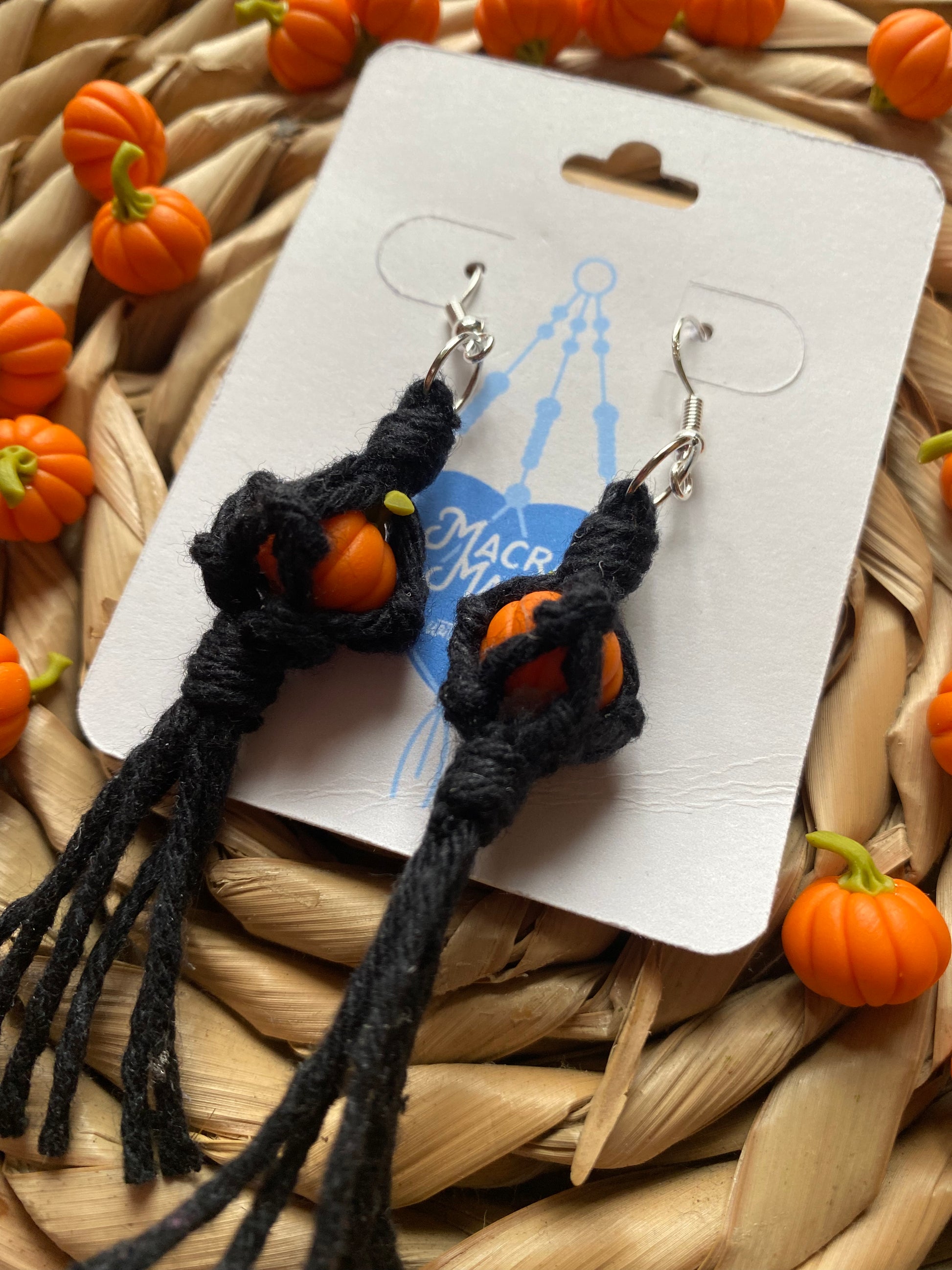 A pair of earrings made with small orange pumpkins wrapped in black yarn on silver hooks, displayed on a white backing card with a wicker mat and lots of small orange pumpkins in the background.  