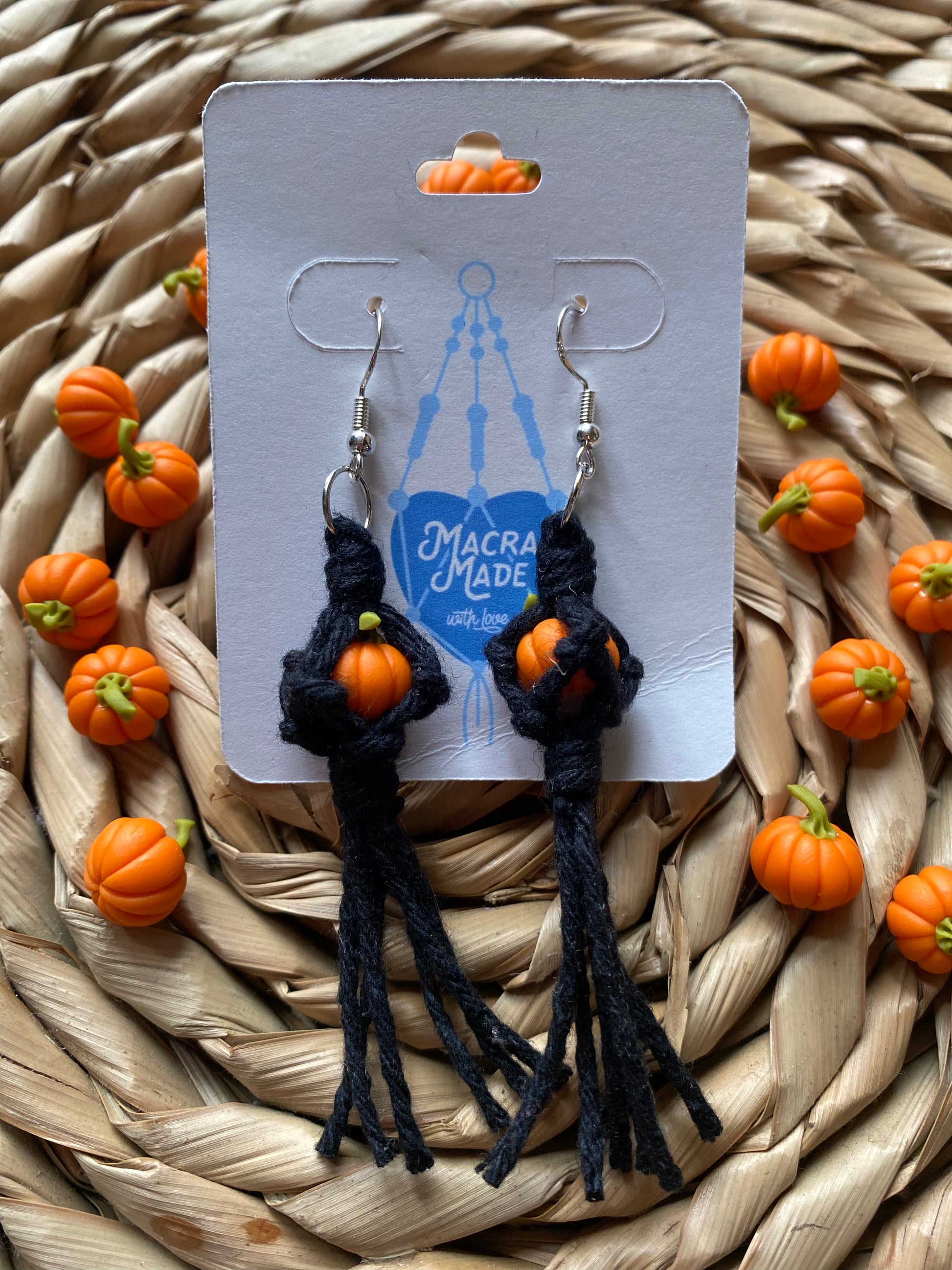 A pair of earrings made with small orange pumpkins wrapped in black yarn on silver hooks, displayed on a white backing card with a wicker mat and lots of small orange pumpkins in the background.  