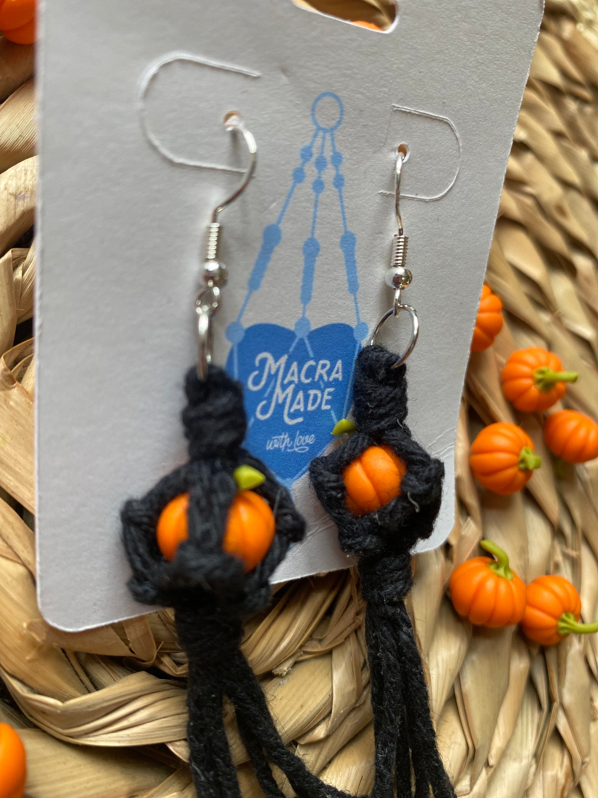 Side angle of a pair of earrings made with small orange pumpkins wrapped in black yarn on silver hooks, displayed on a white backing card.