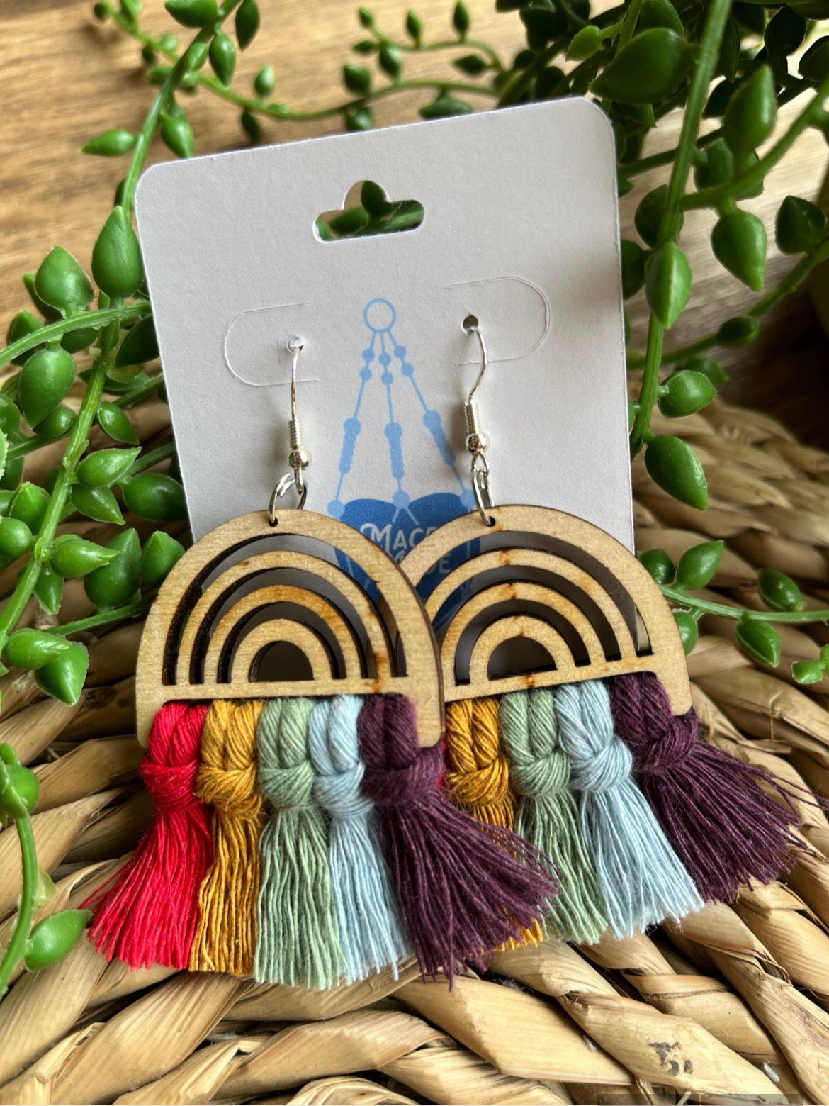 Wooden earrings with colourful tassels on a white card, surrounded by greenery.