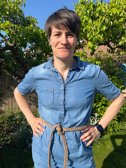 A person stands in a garden with green grass and plants in the background. They are wearing a denim dress with a macrame belt made from beige recycled cotton braided yarn. 