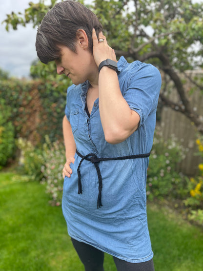 A person wearing a blue denim dress and a black wristwatch, along with a braided Macrame Belt stands in a garden, one hand on the back of their neck and the other on their hip. The background features green grass, assorted flowers, and a tree.