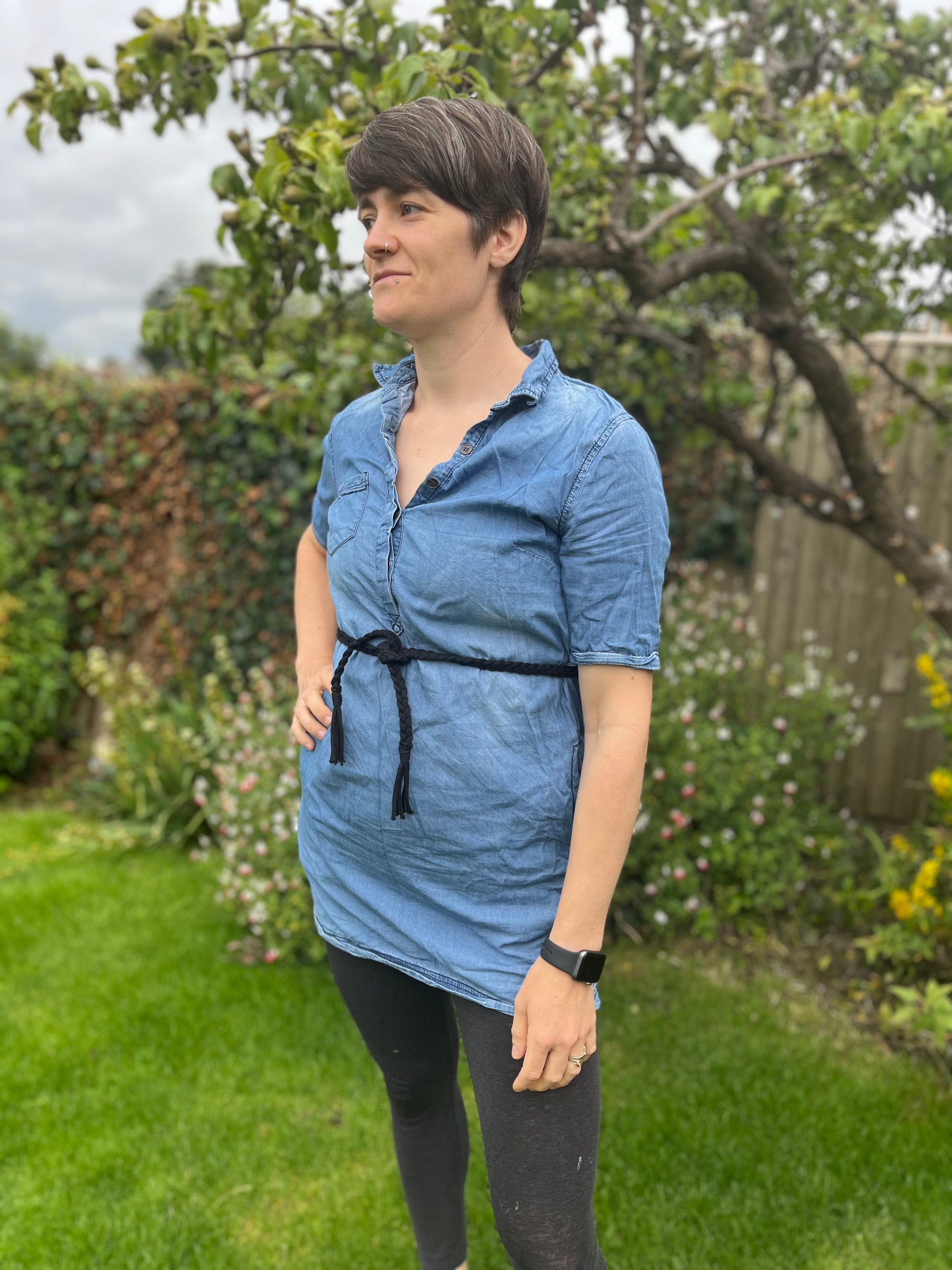 A person with short hair stands in a garden wearing a blue button-up shirt with black leggings and a braided macrame belt made with black yarn. The background features greenery, flowers, and a tree.