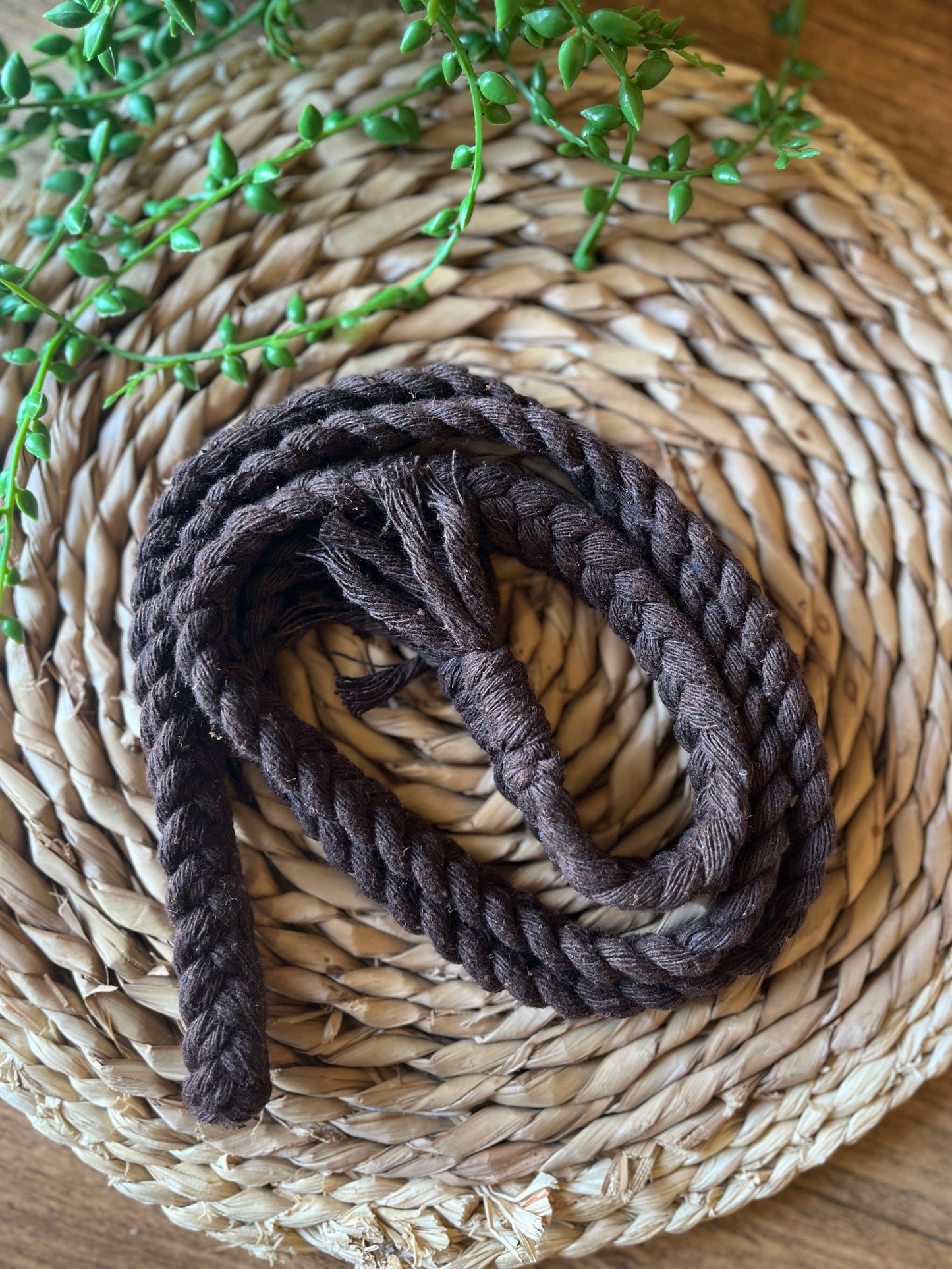 A coiled brown cotton braided macrame rope belt with tasseled ends, rests on a round wicker surface. 