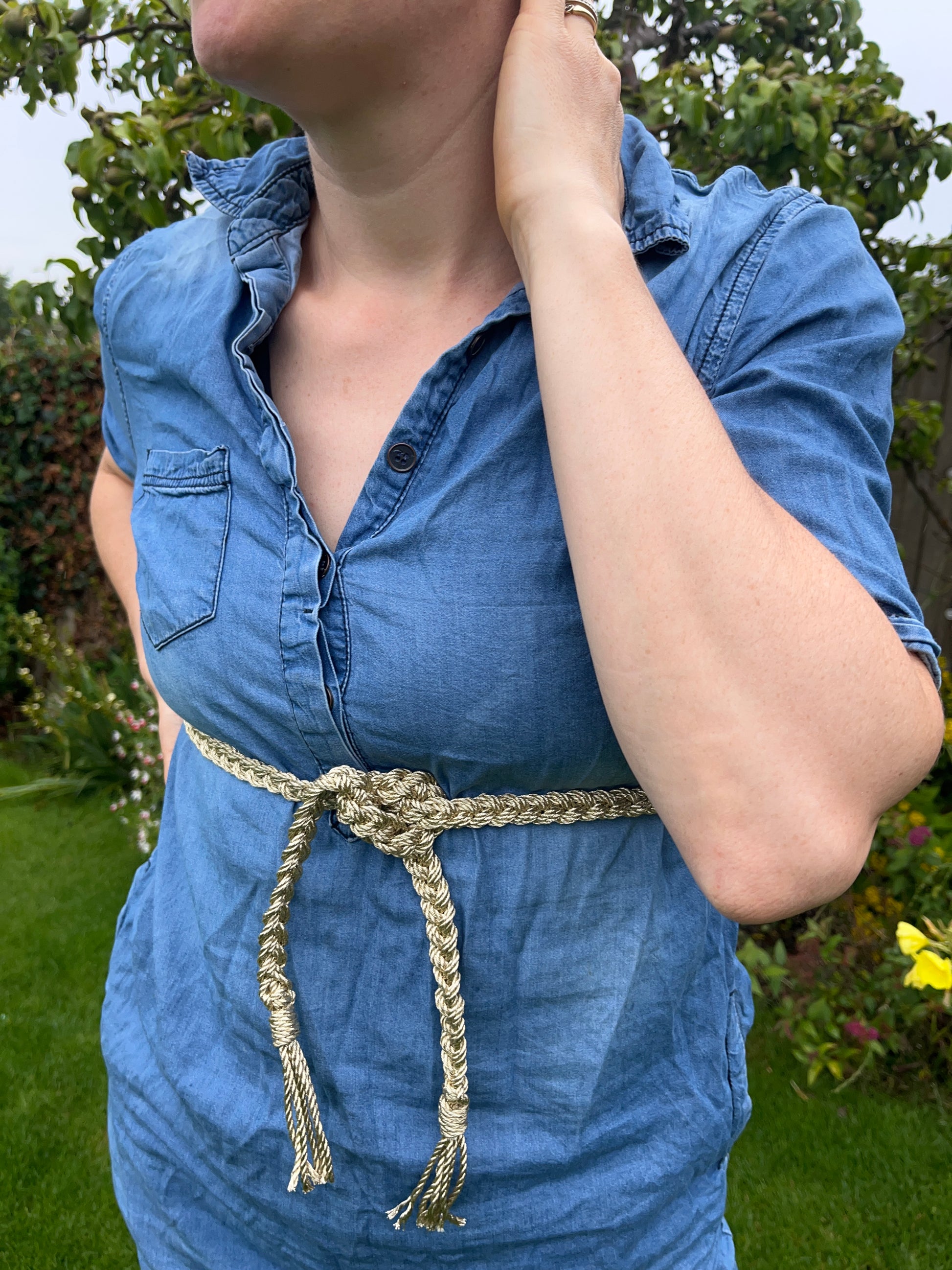 A person wearing a blue denim dress and a braided gold macrame belt stands in a garden, one hand on the back of their neck and the other on their hip. The background features green grass, assorted flowers, and a tree.
