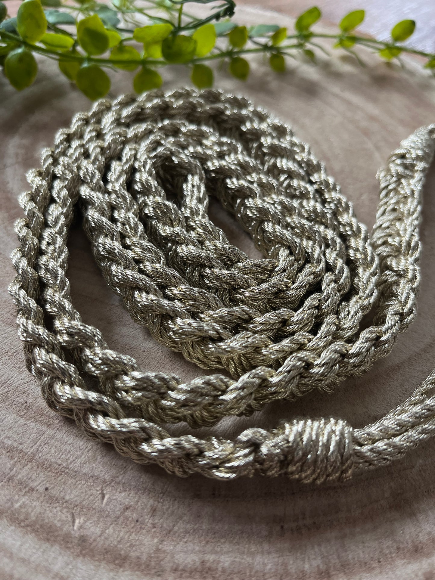A coiled gold braided rope belt rests on a round wooden surface. 