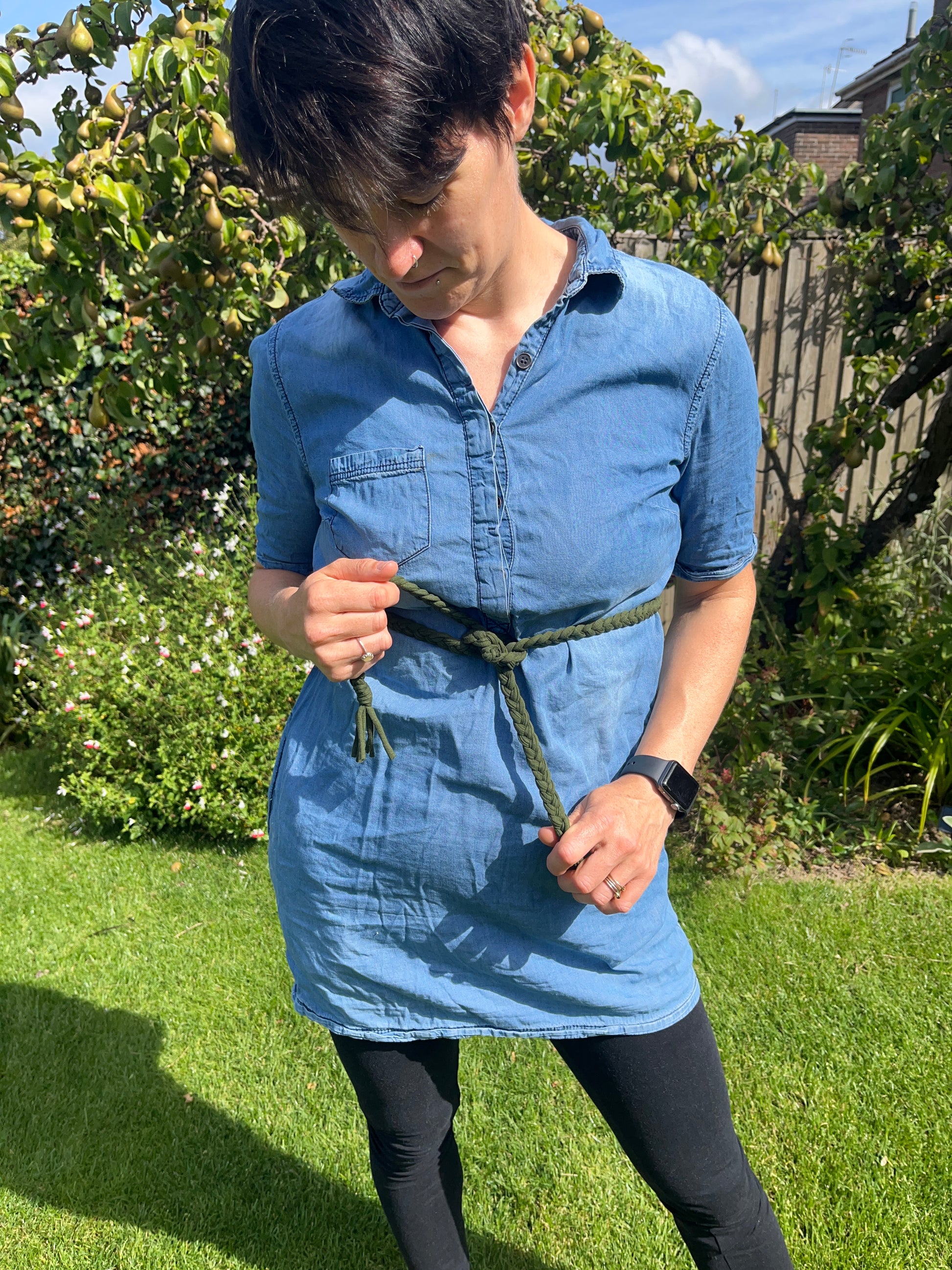 A person wearing a blue denim tunic dress and a green macrame belt stands in a garden, looking down, whilst holding the ends of the belt. The background features green grass, assorted flowers, and a tree.