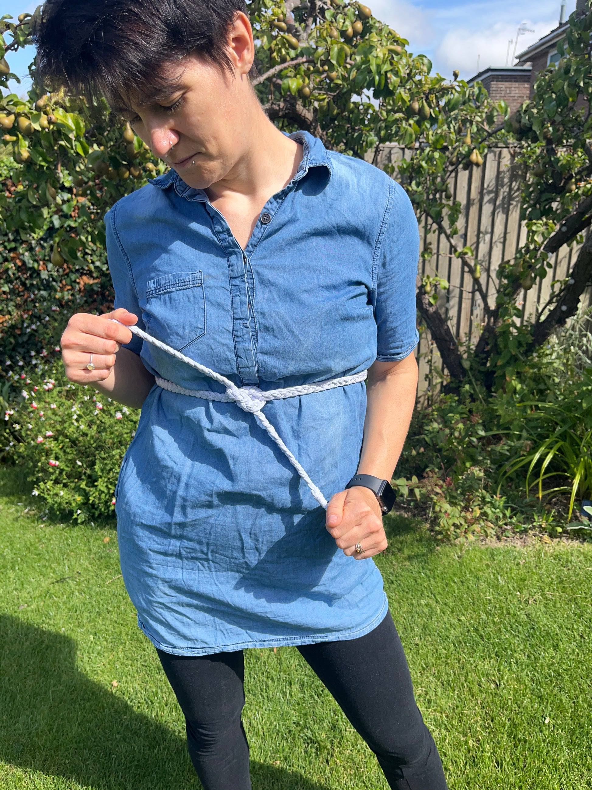 A person wearing a blue denim dress and a grey macrame belt stands in a garden, looking down, whilst holding the ends of the belt. The background features green grass, assorted flowers, and a tree.