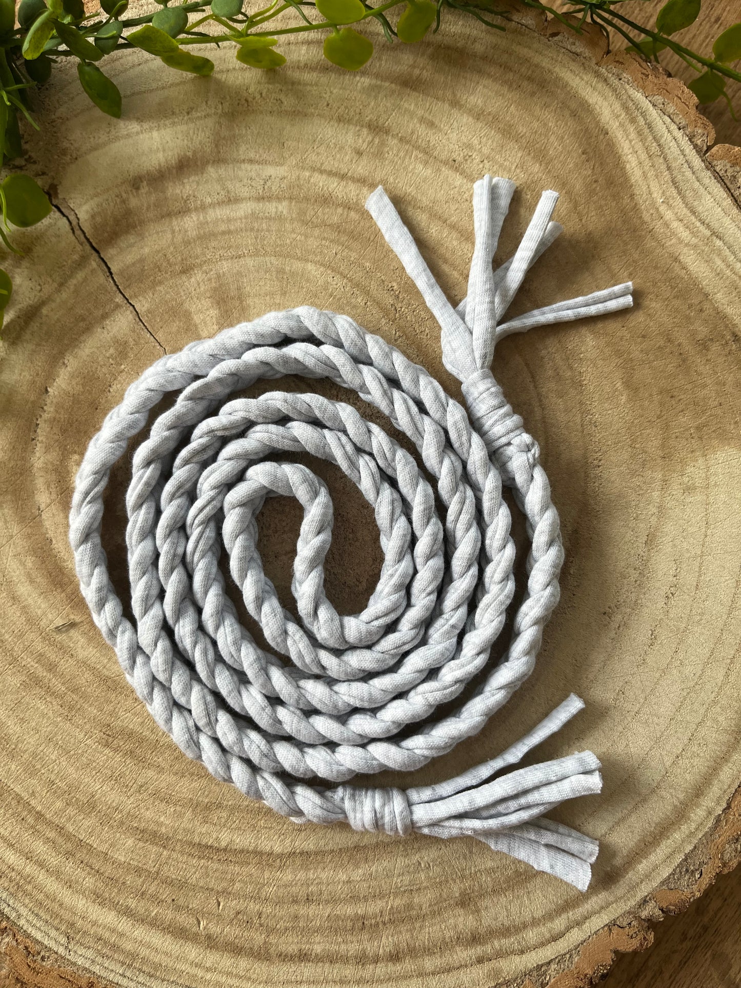 A coiled grey t shirt yarn belt with tasseled ends, rests on a round wooden surface. 
