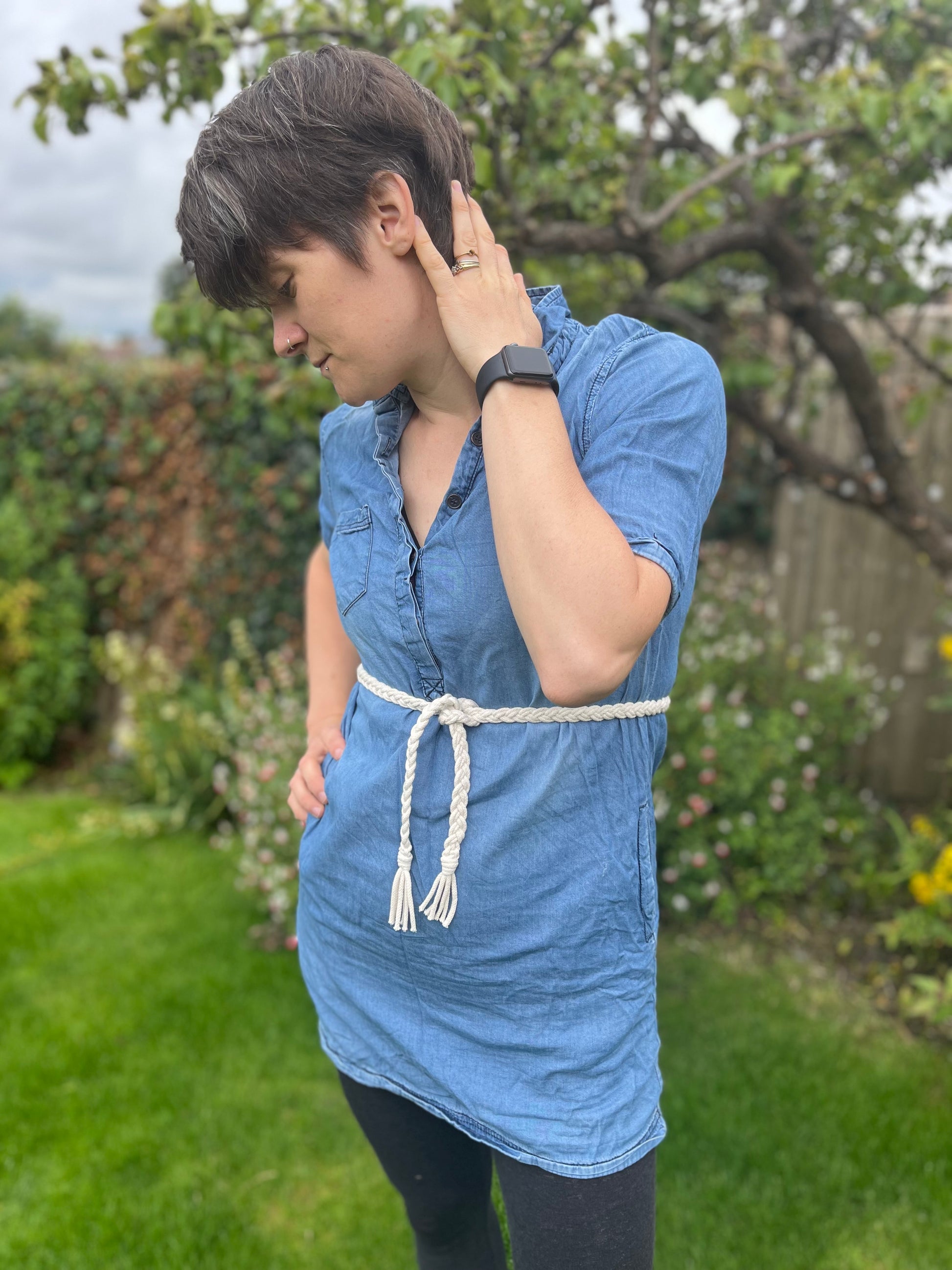 Person with short hair stands in a garden, wearing a blue denim tunic over black leggings. A braided macrame belt is tied around their waist. They are looking down and have one hand touching their neck and the other resting on their hip. Trees and plants are in the background.