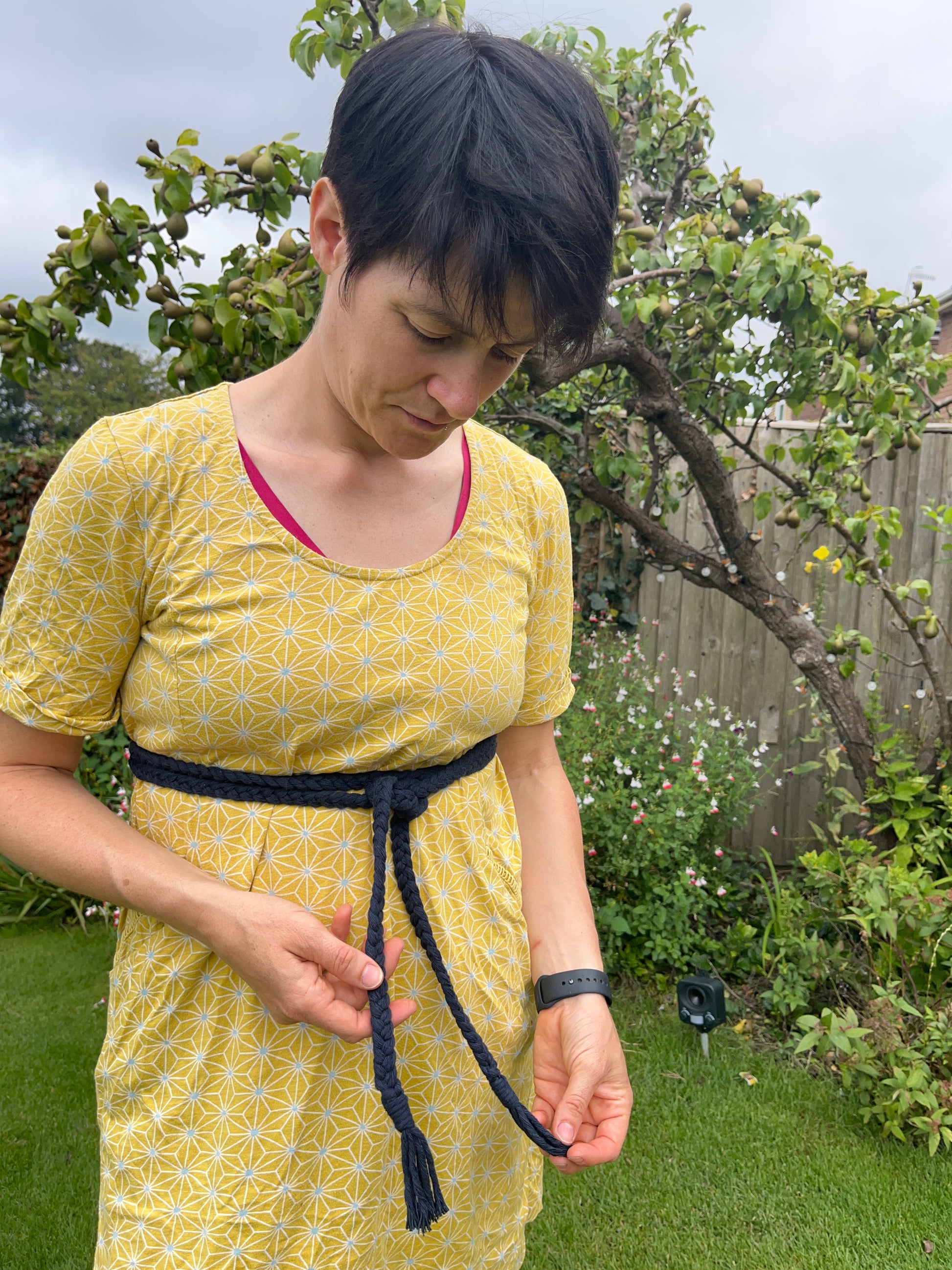 A person wearing a yellow dress and a navy macrame belt stands in a garden, looking down, whilst holding the ends of the belt. The background features green grass, assorted flowers, and a tree.