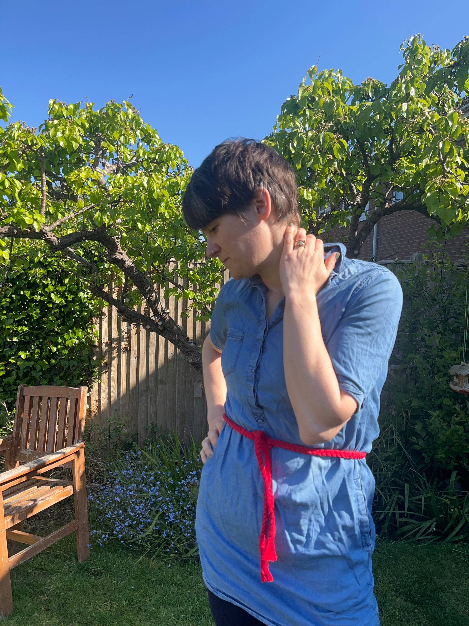 Person with short hair stands in a garden, wearing a blue denim tunic over black leggings. A red braided macrame belt is tied around their waist. They are looking down and have one hand touching their neck and the other resting on their hip. Trees and plants are in the background.