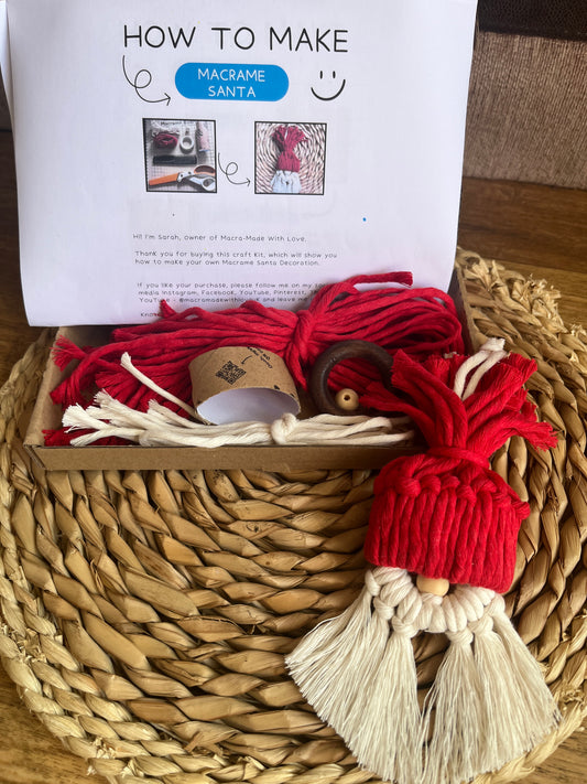 Macrame santa decoration placed on a wicker mat in front of an open box containing DIY craft materials and instructions for a macramé christmas craft kit. 