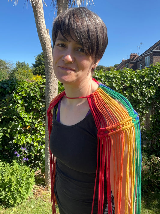 A female wearing a black vest, and handmade macrame shoulder accessory epaulette, stands outside on a sunny day, smiling towards the camera. 