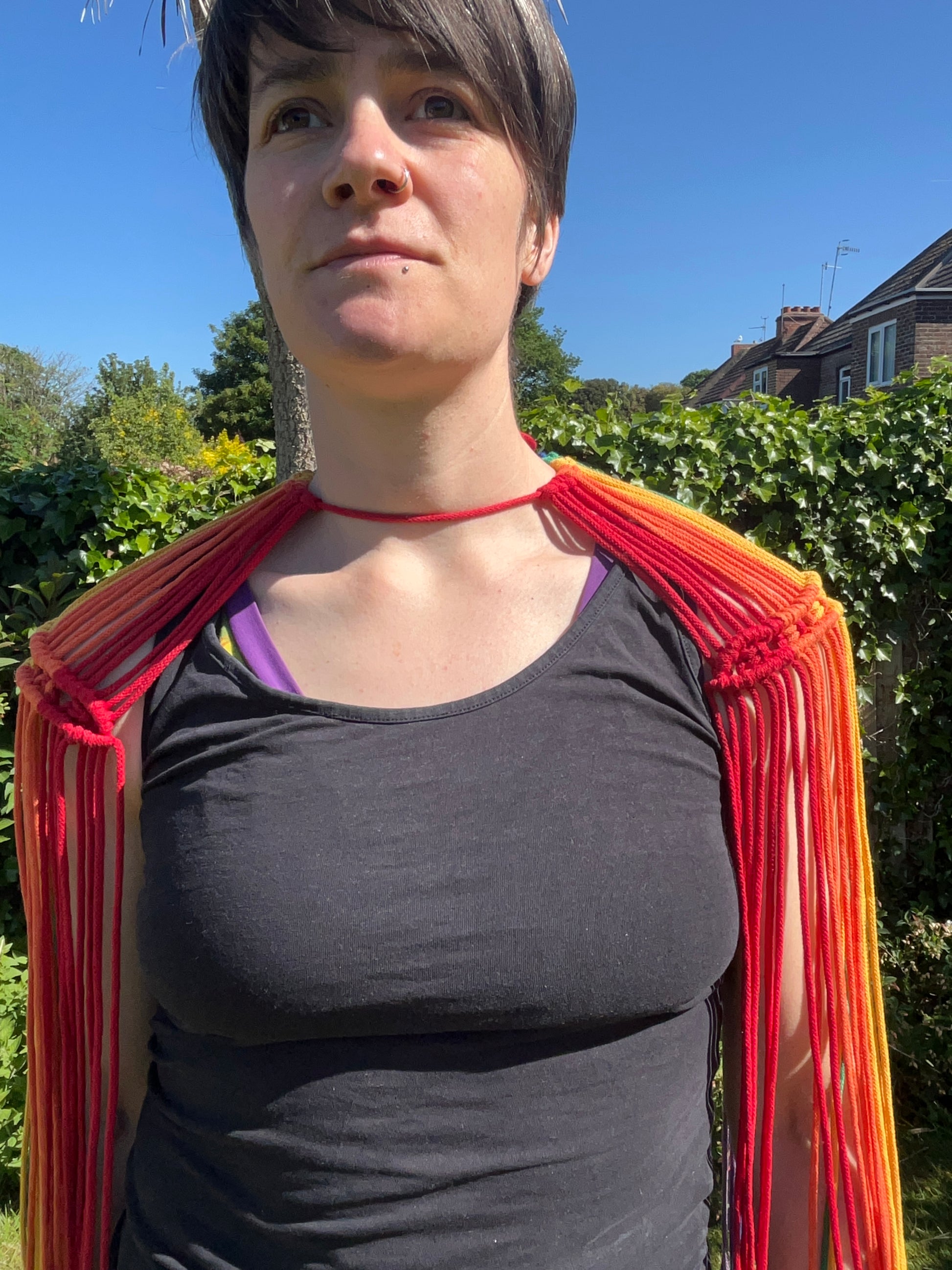 A female wearing a black vest, and handmade macrame shoulder accessory epaulette, stands outside on a sunny day, smiling towards the camera. 
