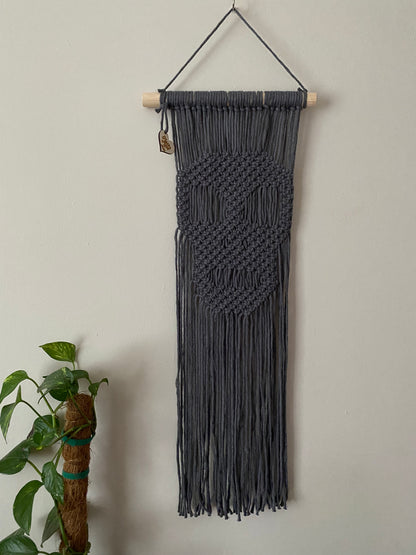 Long dark grey macrame wall hanging with a knotted pattern in the shape of a skull, made on a wooden dowel, hanging against a beige wall next to a tall green houseplant. 