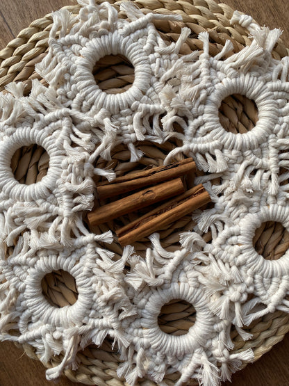 Six handmade macrame snowflake ornaments, knotted with natural coloured cotton yarn, displayed in a circle on a wicker surface with cinnamon sticks in the middle. 
